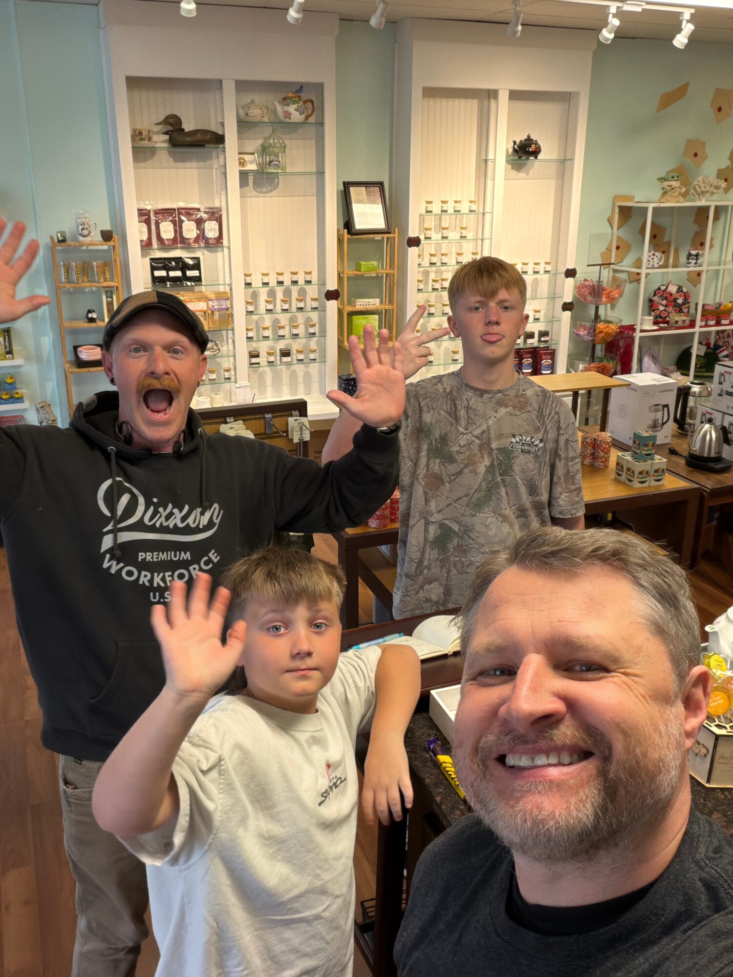 Four people inside a store, smiling and waving, shelves with products in the background.