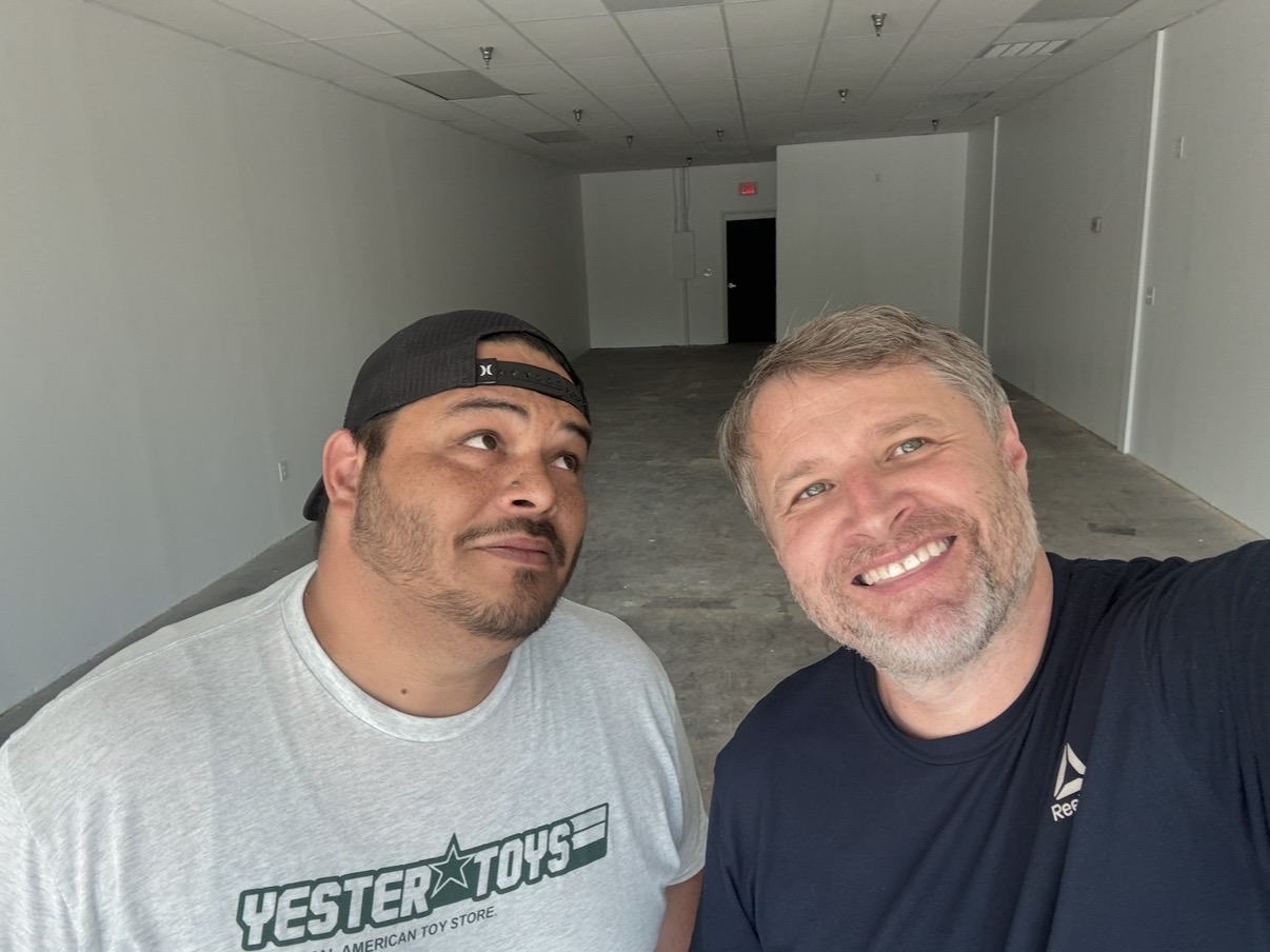 Two men smiling, one with a beard and the other wearing a cap, in an empty hallway.