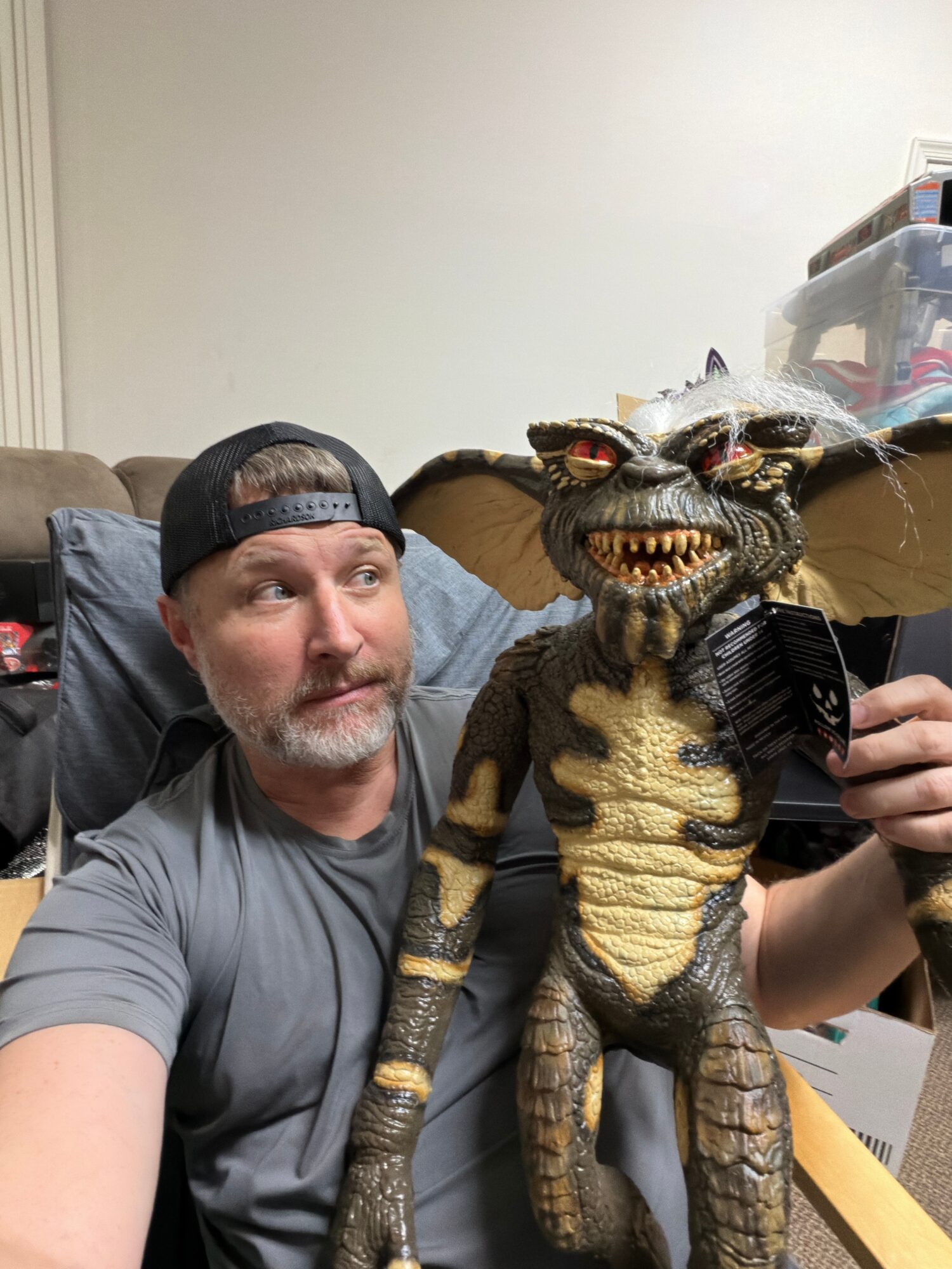 Man with a beard and cap looking at a detailed dragon puppet with wings, sharp teeth, and textured skin, held by a person.