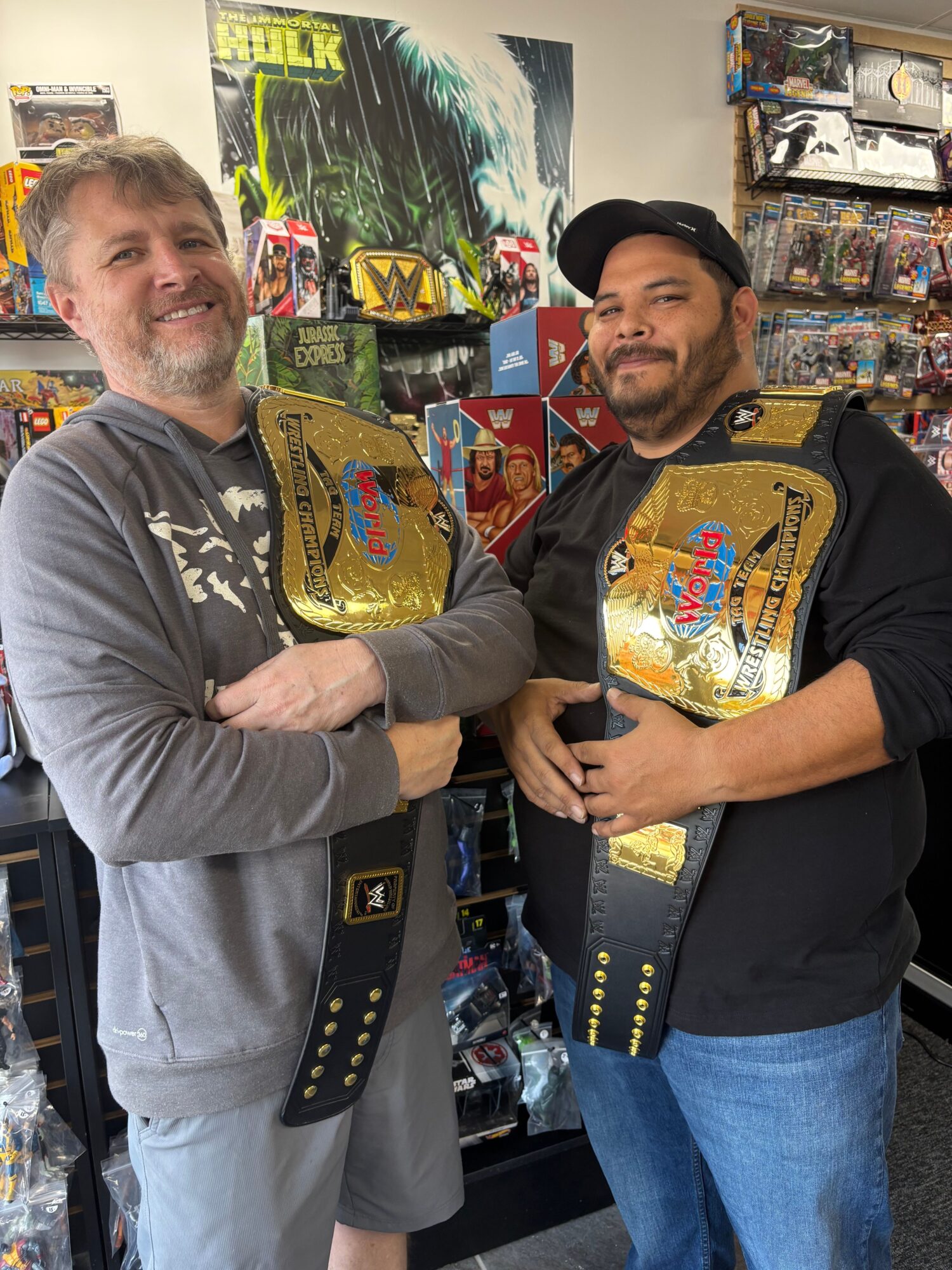 Two men holding championship belts in a store, with posters and merchandise in the background.