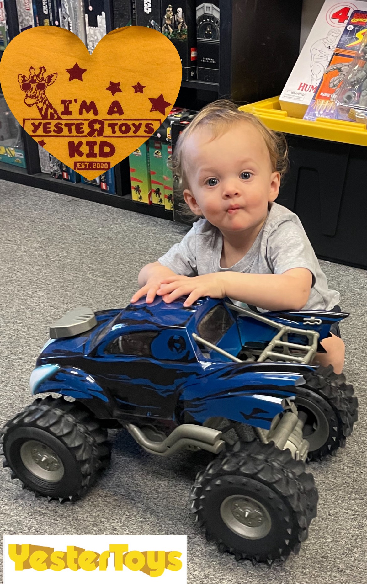 Child sitting on floor with a blue toy monster truck, holding it, in a store aisle with toys and a yellow heart-shaped sign.