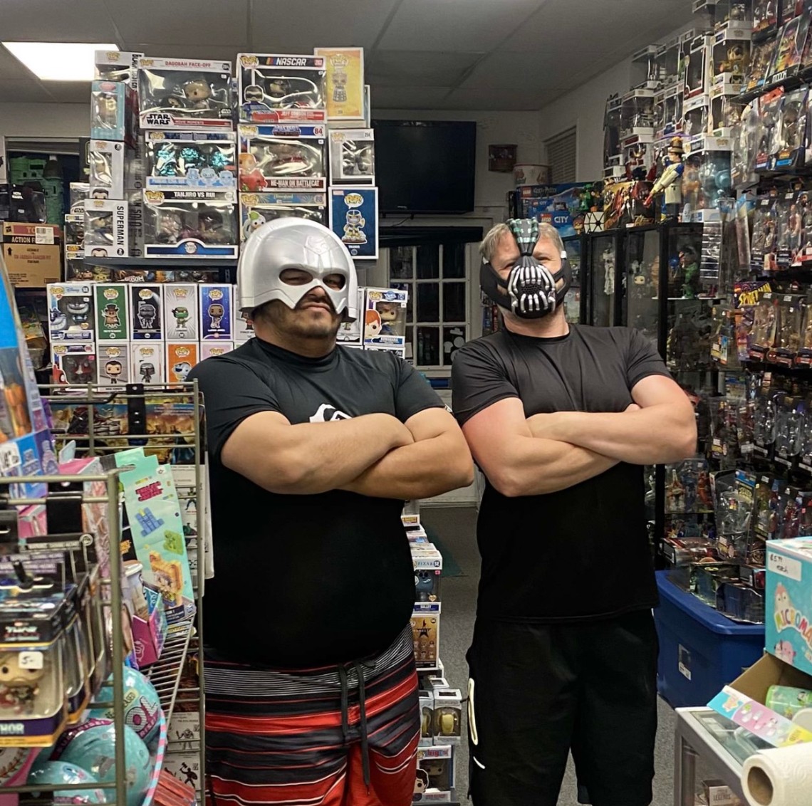 Two people wearing masks and black shirts with arms crossed inside a toy store, shelves filled with toys and collectibles behind them.