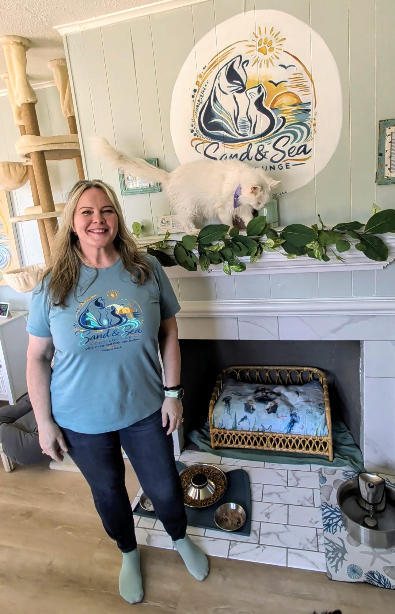 Woman with long hair smiling, wearing a blue t-shirt with a swan logo, standing in front of a fireplace with a swan-themed sign and decorations.