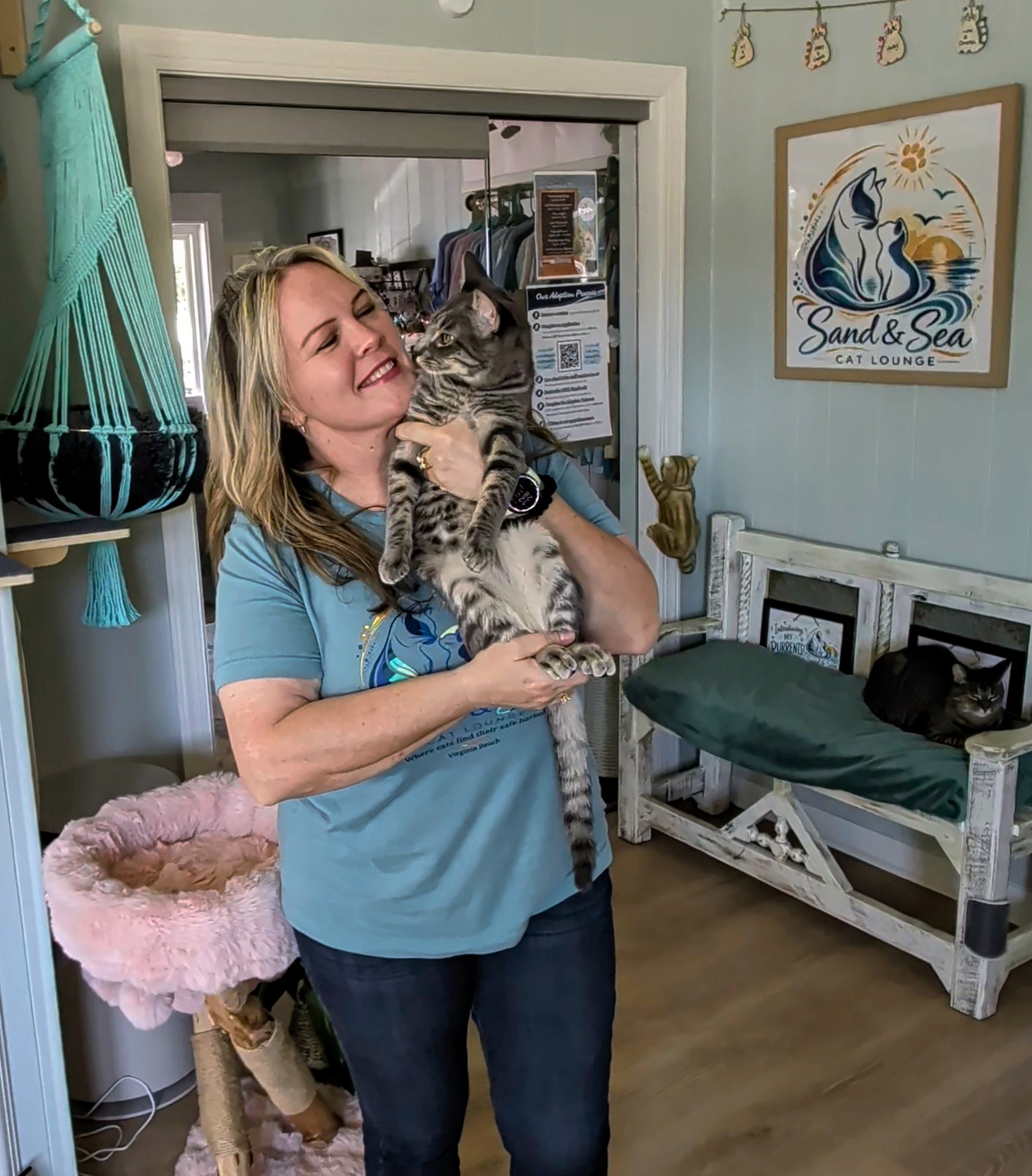 Woman holding a cat indoors, smiling, with a beach-themed picture on the wall, a bench, and a pink cat tree nearby.