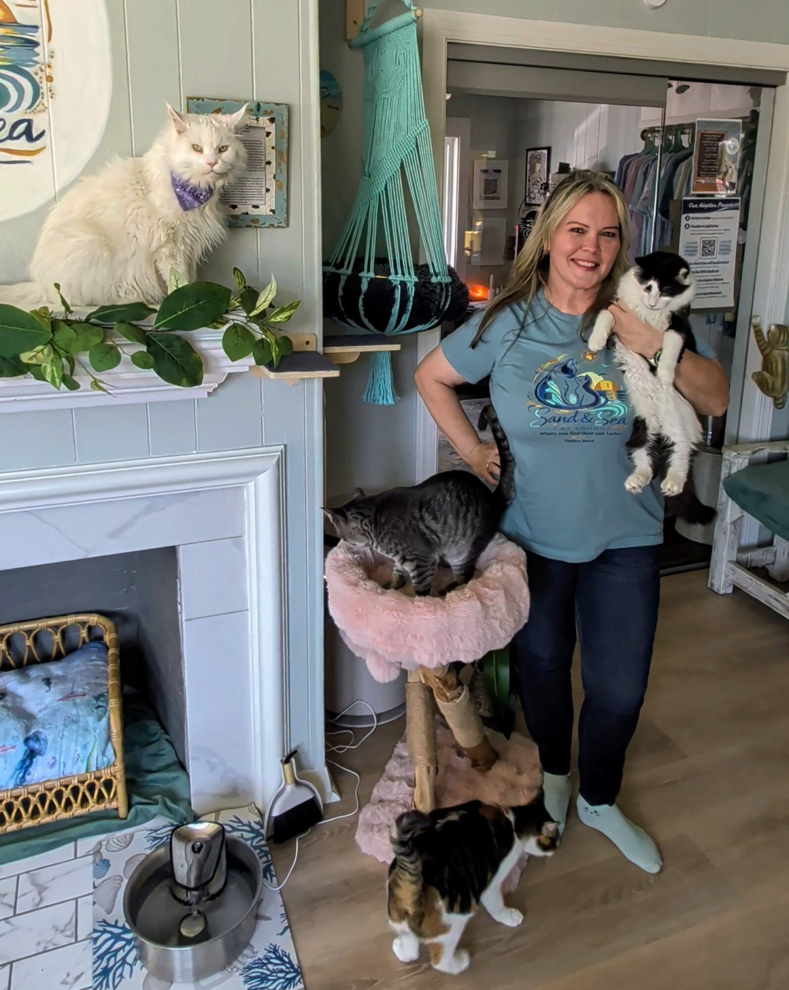 Woman holding a black and white cat, standing near a fireplace with a white cat on the mantel, in a cozy room.