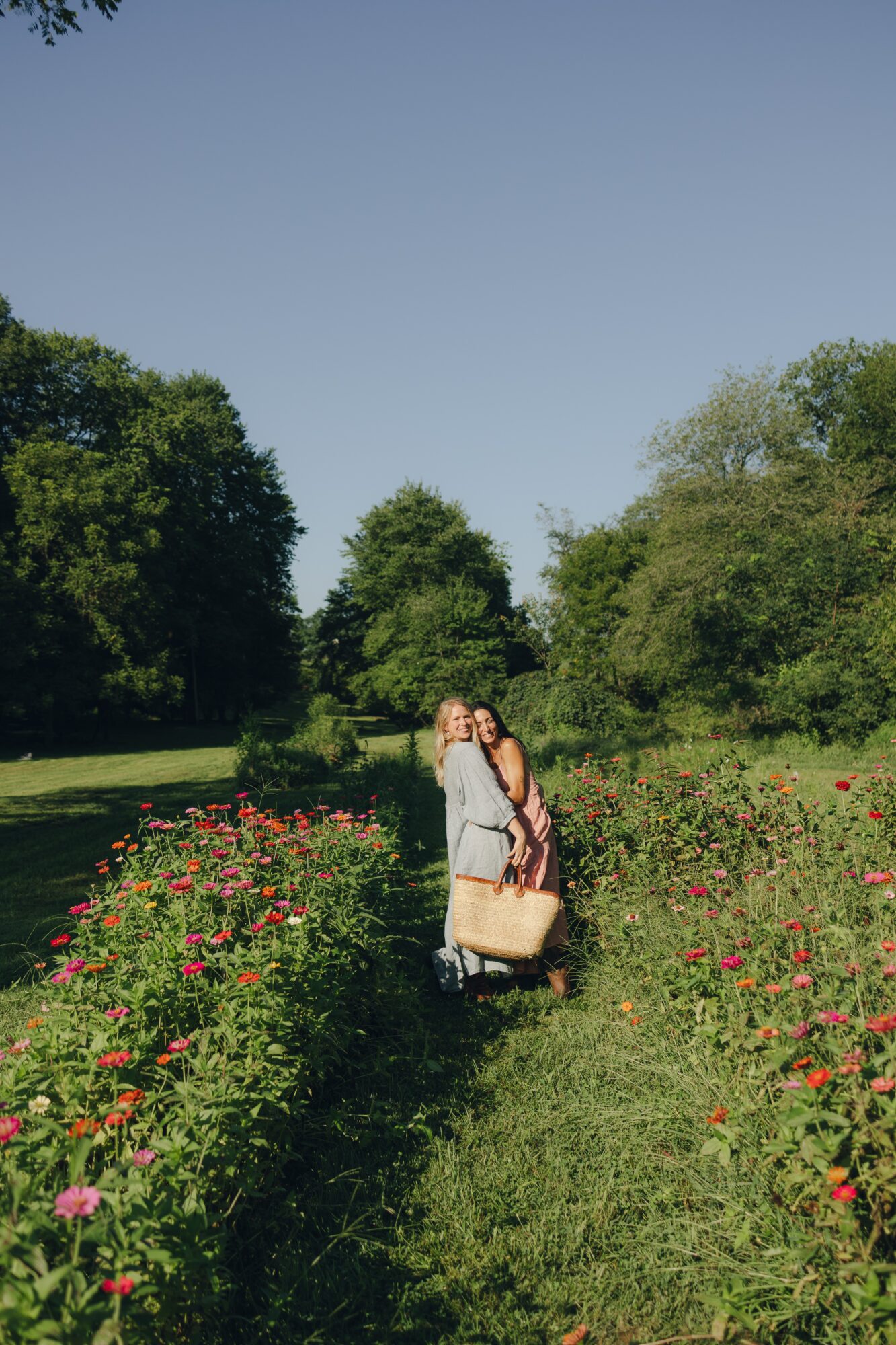 Two people walking along a garden path surrounded by flowering bushes and green trees under a clear sky.