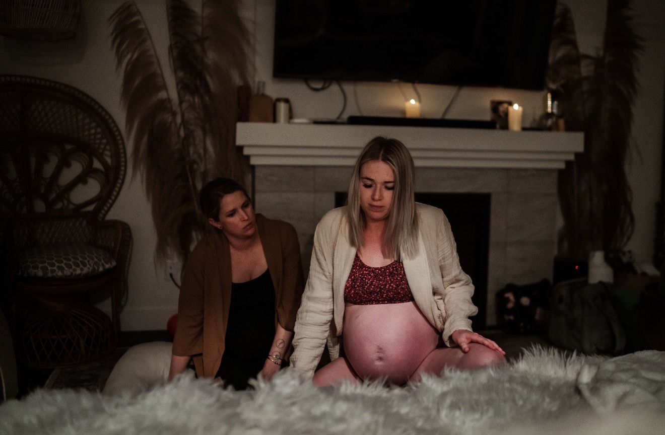 Two women sit on a carpeted floor in a living room, one pregnant, with a fireplace and TV behind them.
