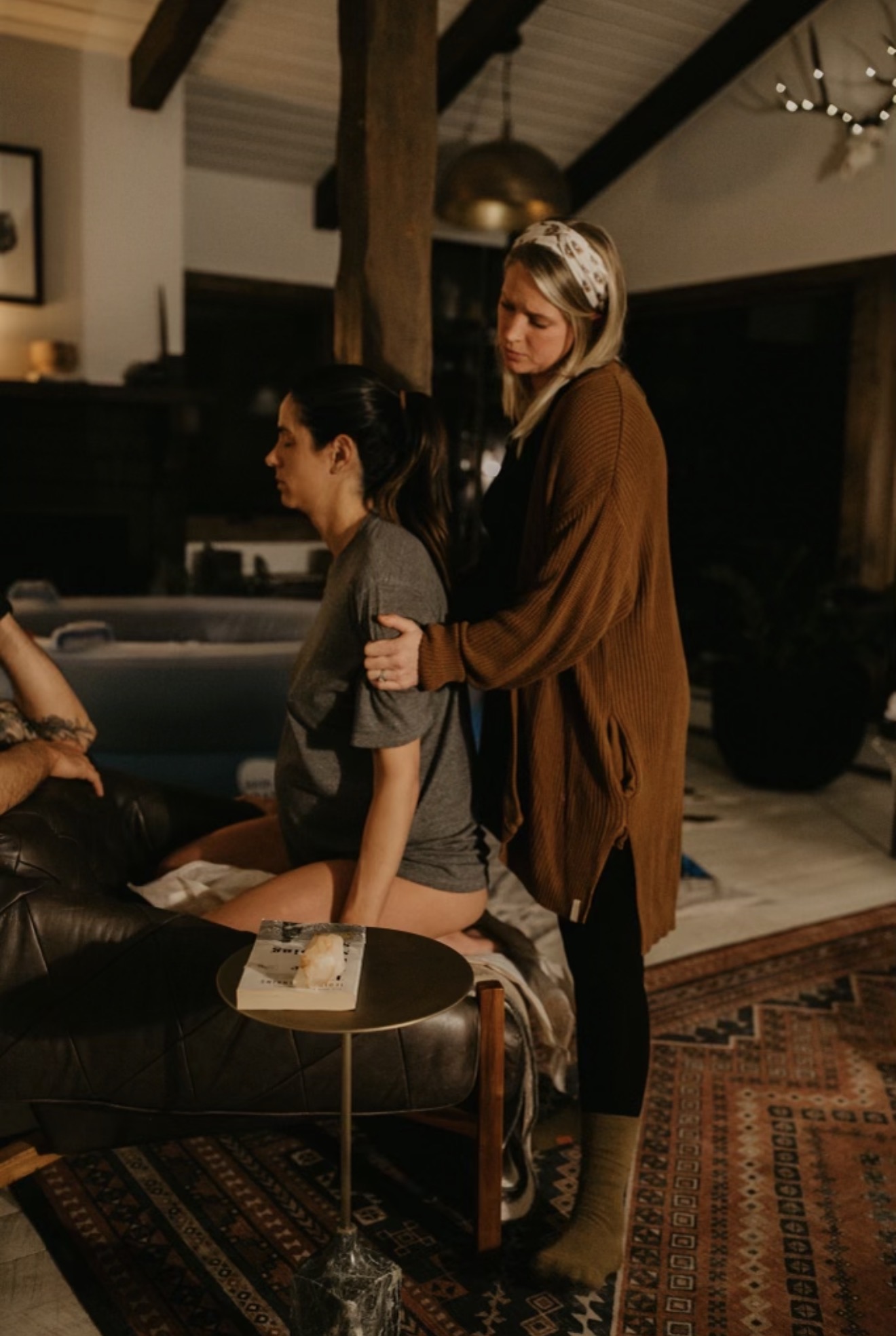 Two women in a cozy room, one standing and the other sitting, with warm lighting and a patterned rug.