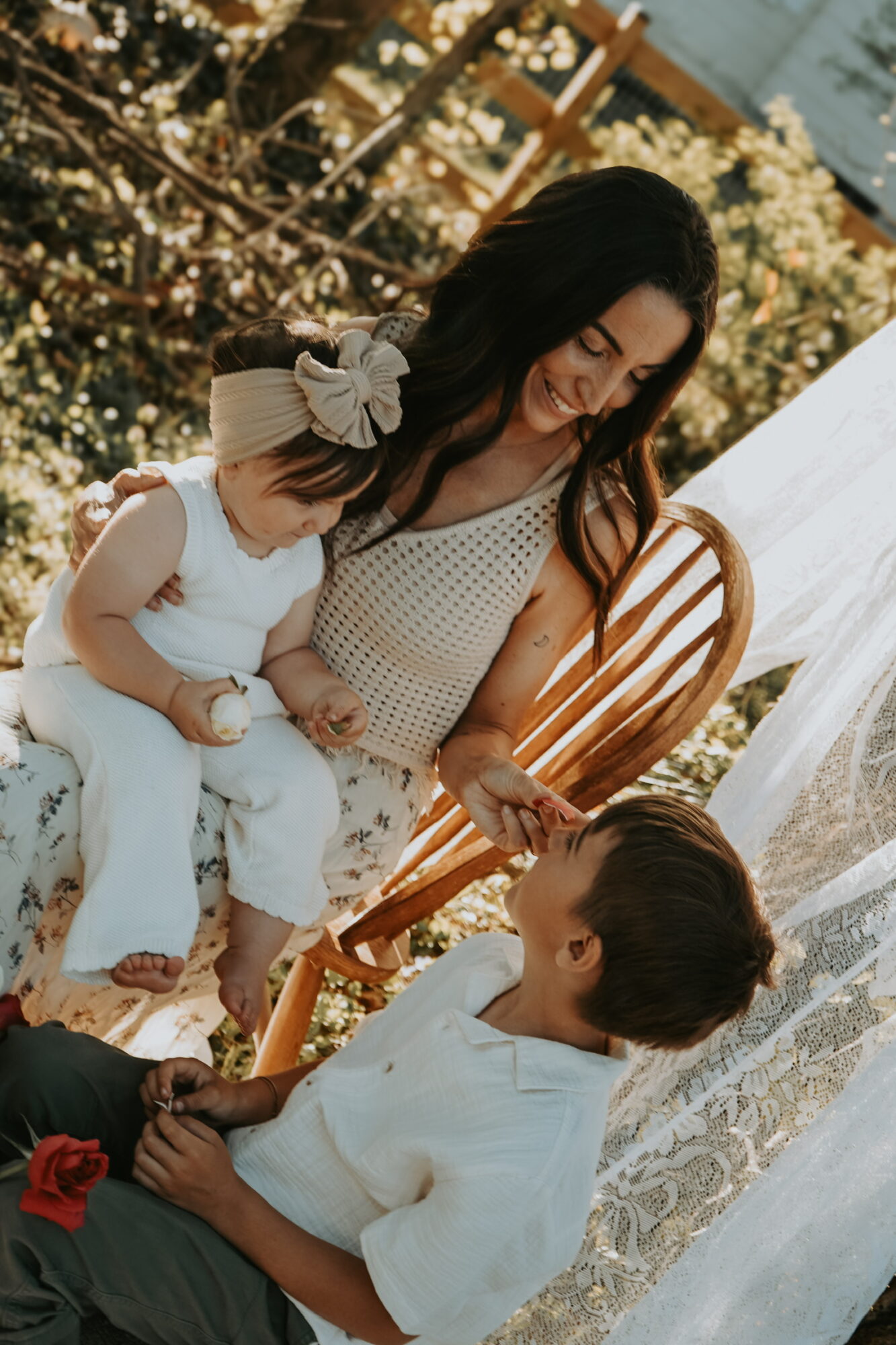 Woman with two children outdoors, one girl with a bow in her hair and a boy, sitting on a wooden chair, smiling.