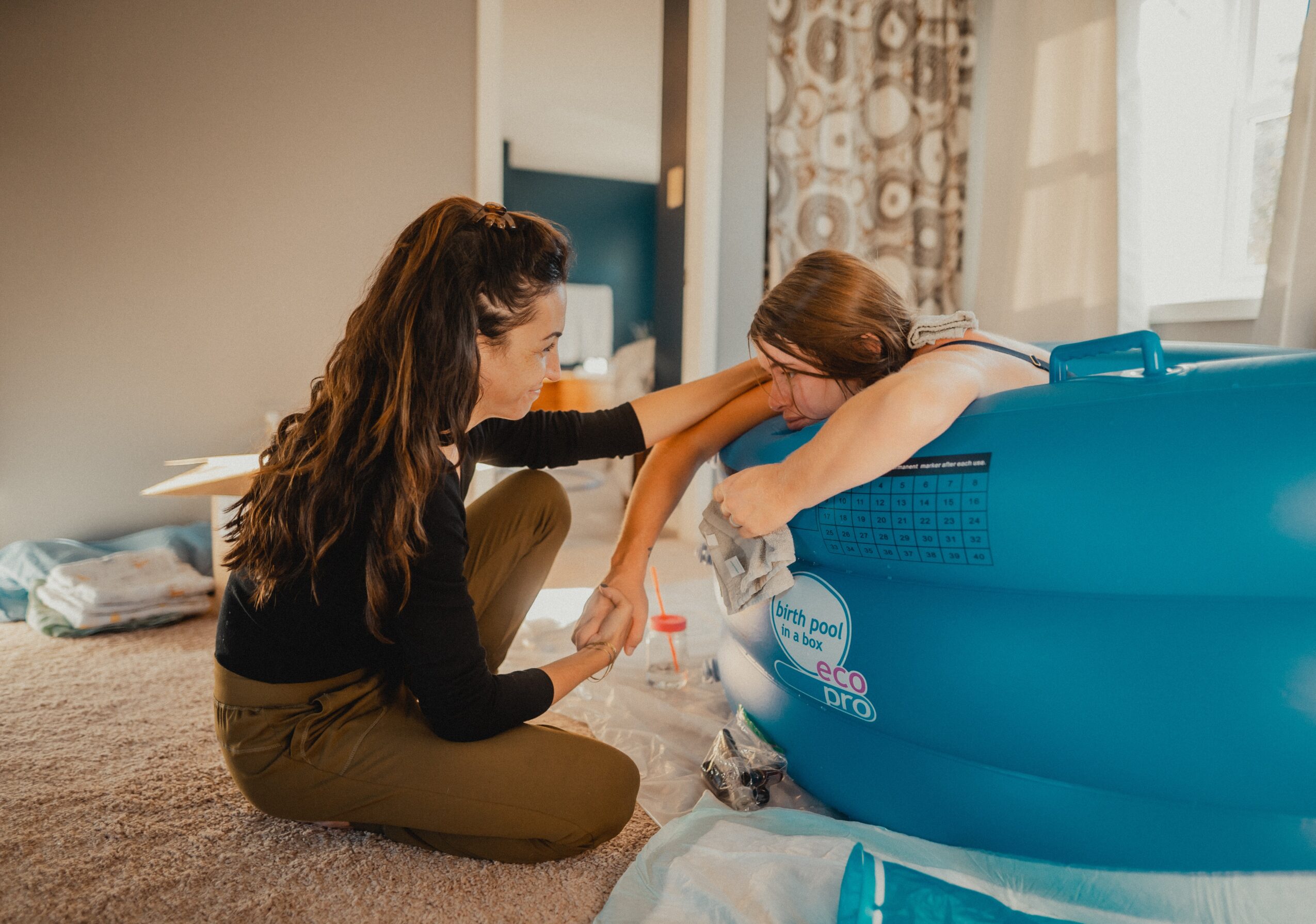 Two women preparing an inflatable pool indoors, one sitting on the floor and the other leaning over it, with a window in the background.