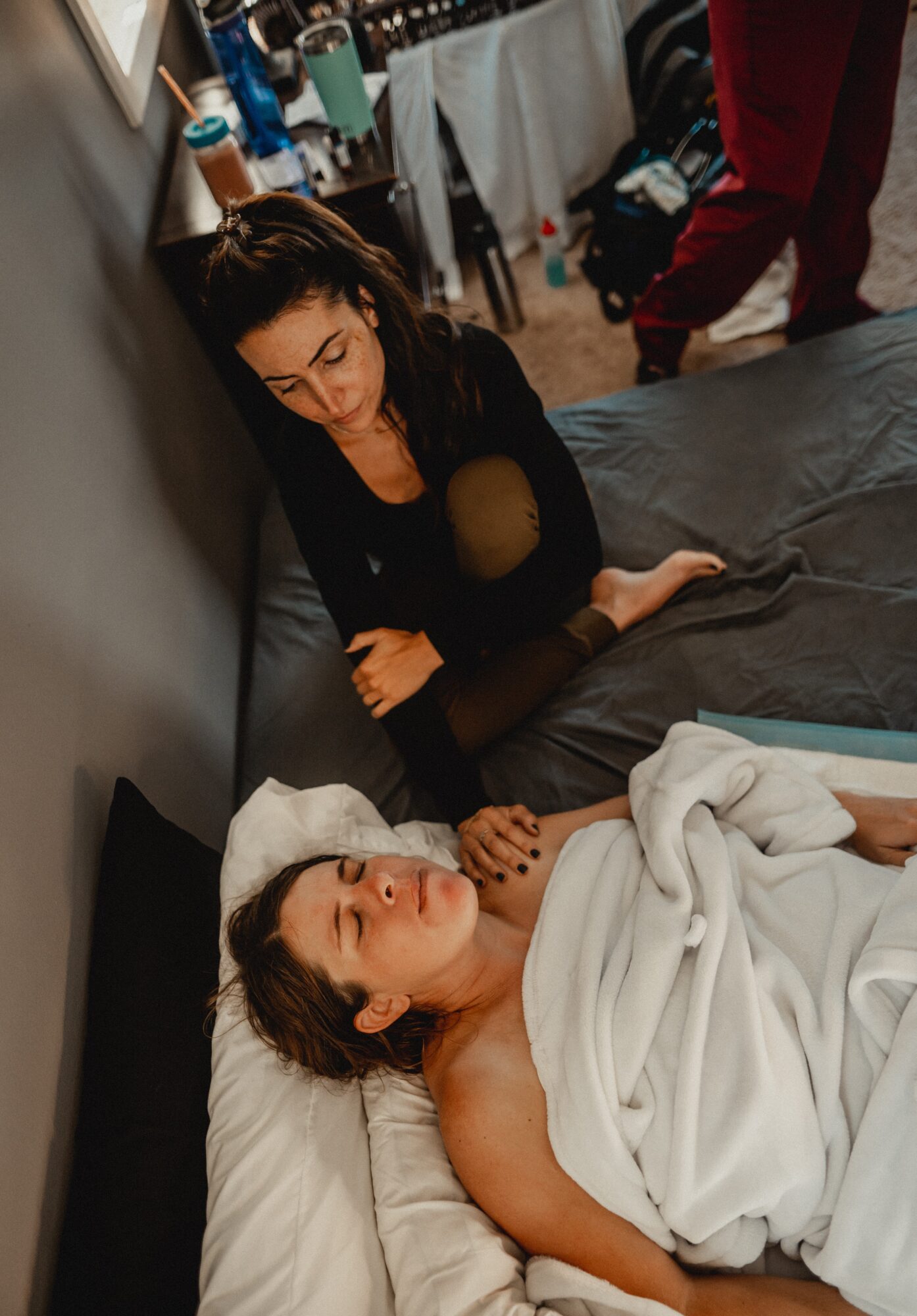 Two women in a room, one lying in bed and the other sitting beside her, with various objects in the background.