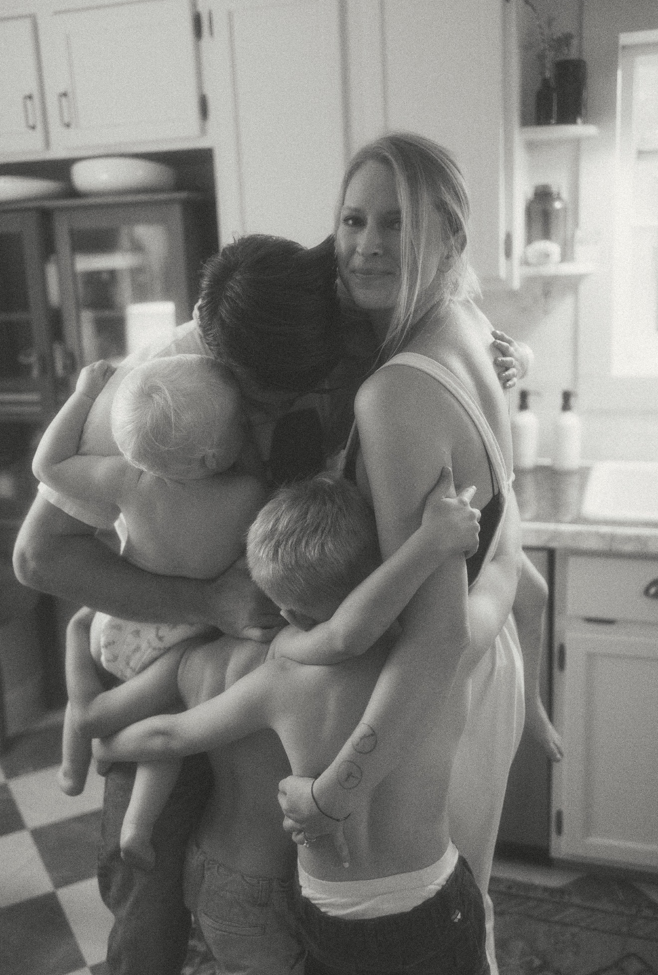 Four people, including a woman and three children, hugging in a kitchen. The woman is smiling at the camera.