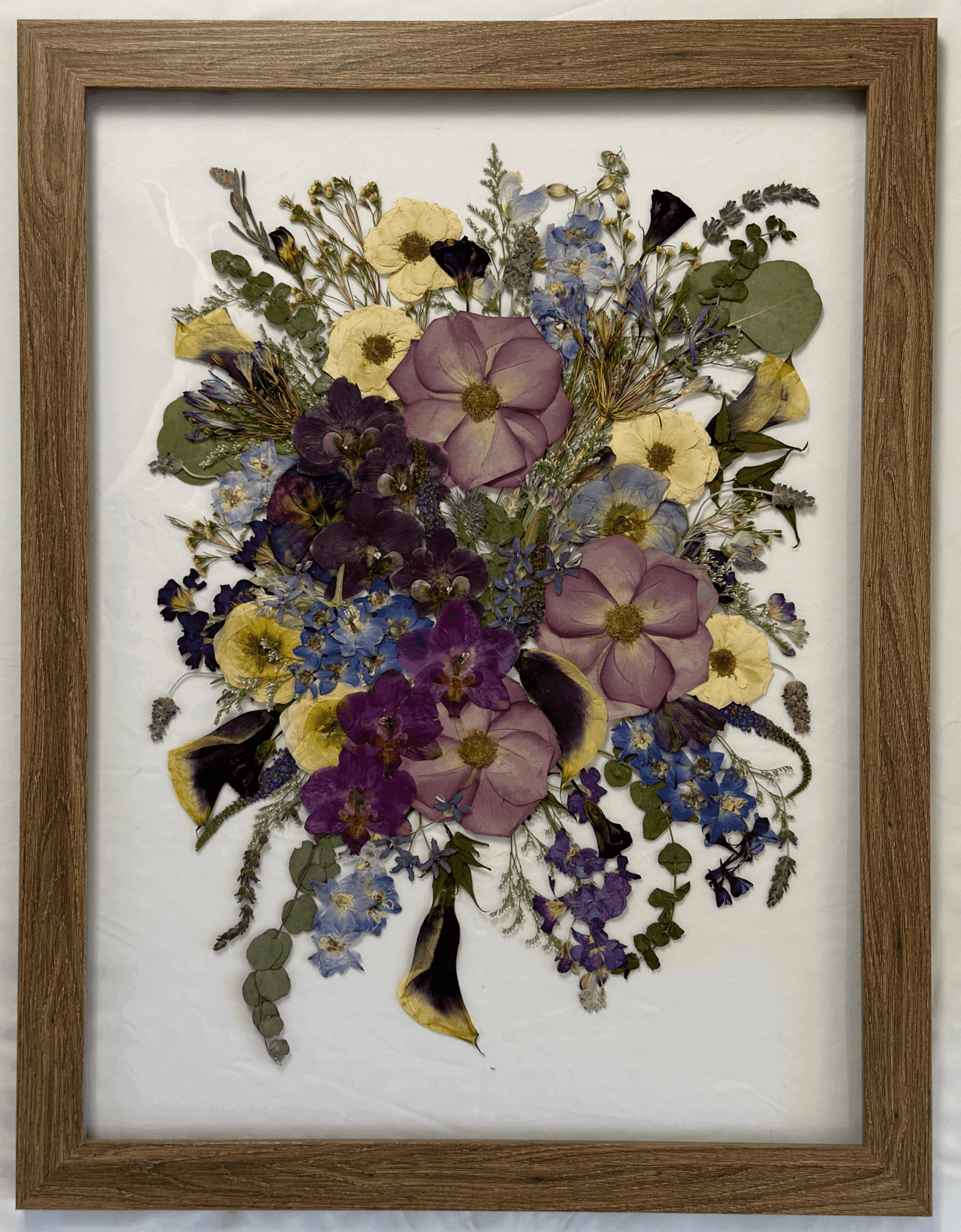 Framed pressed flower arrangement with purple, yellow, and blue flowers and green leaves, in a wooden frame.