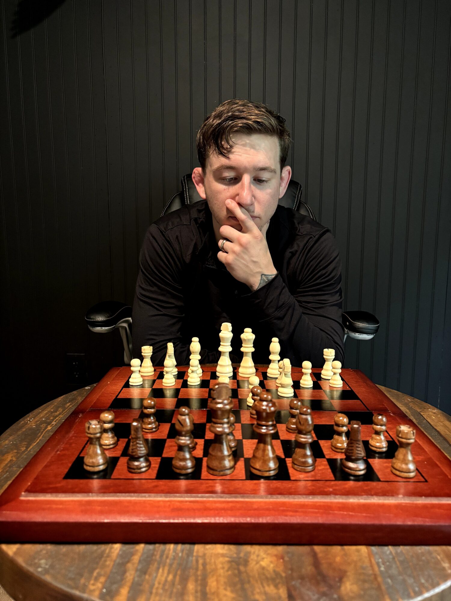 Young man with short hair, wearing a black shirt, concentrating on a chess game against a dark background.