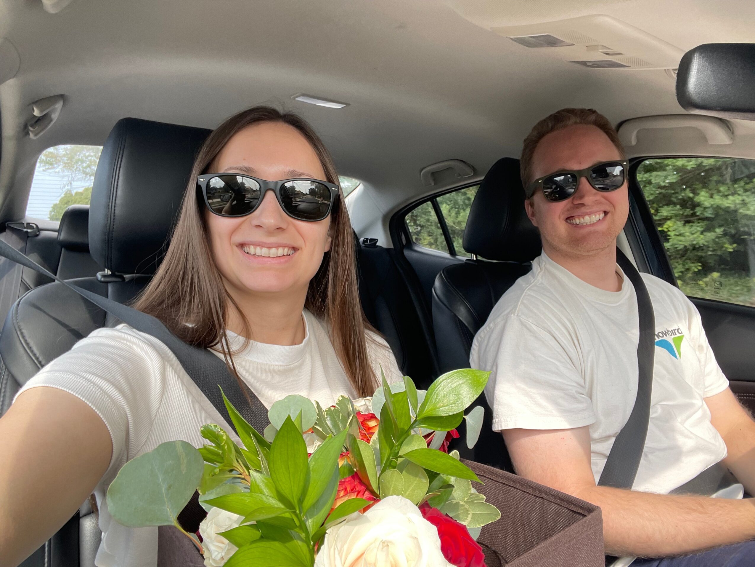 Two women wearing sunglasses sitting in a car, one holding a bouquet of flowers, both smiling.