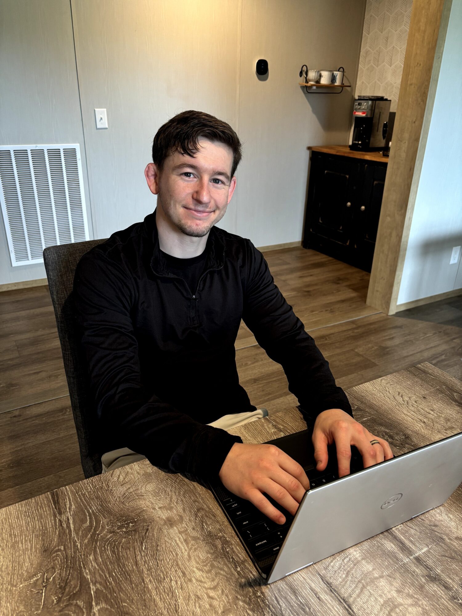 Young man with dark hair and a black shirt sitting at a table using a laptop, in a room with light-colored walls and wooden flooring.