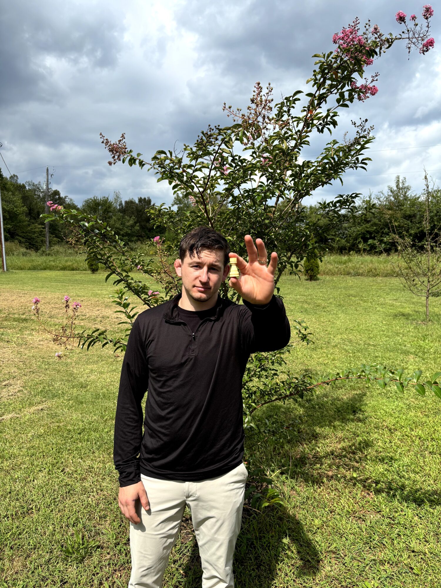 Young man standing outdoors in a grassy field, raising his hand, with a flowering tree and cloudy sky behind him.