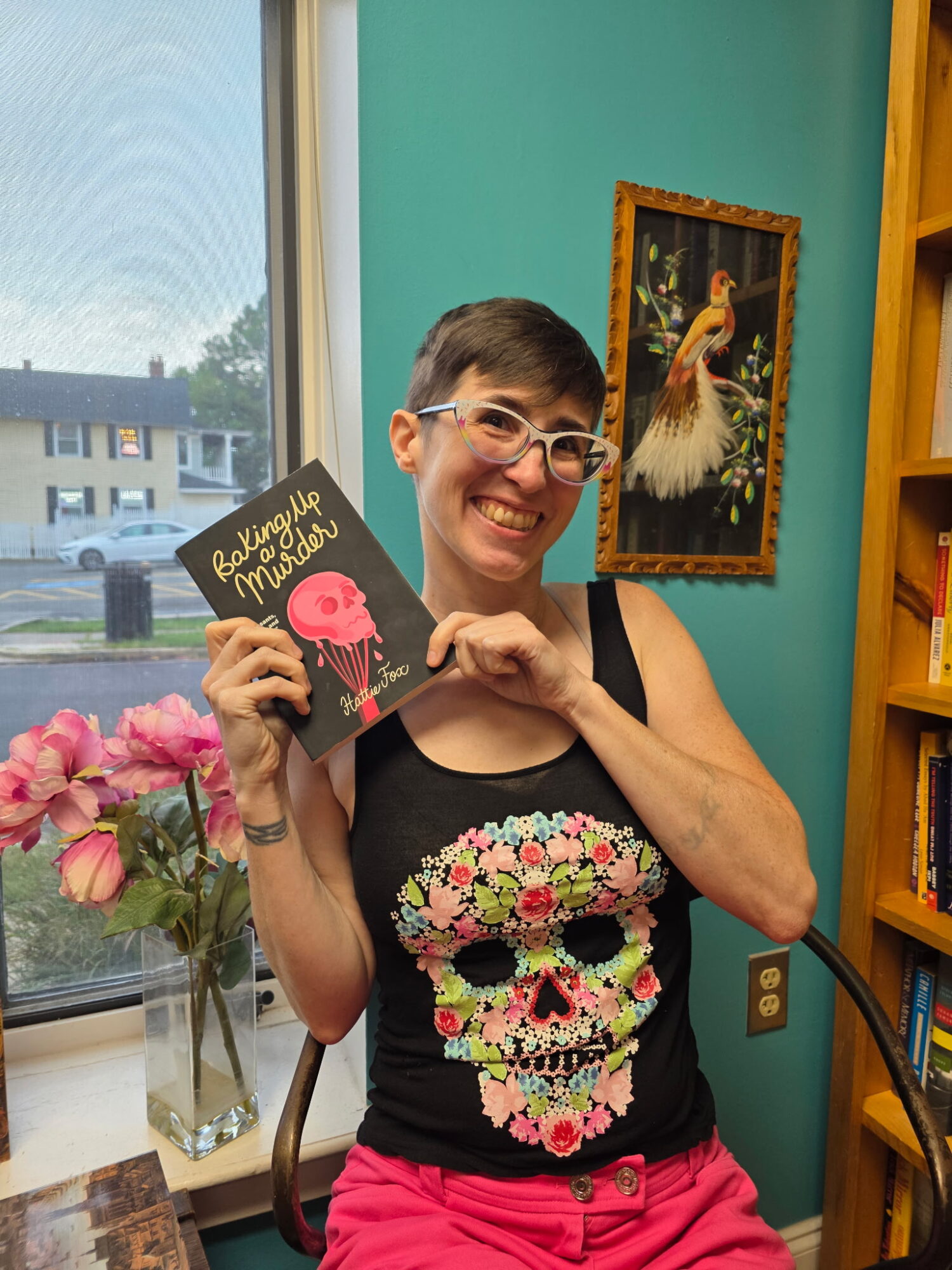 Person with short hair and glasses smiling, holding a book, wearing a black shirt with a colorful skull design, pink pants, standing indoors near a window with pink flowers.