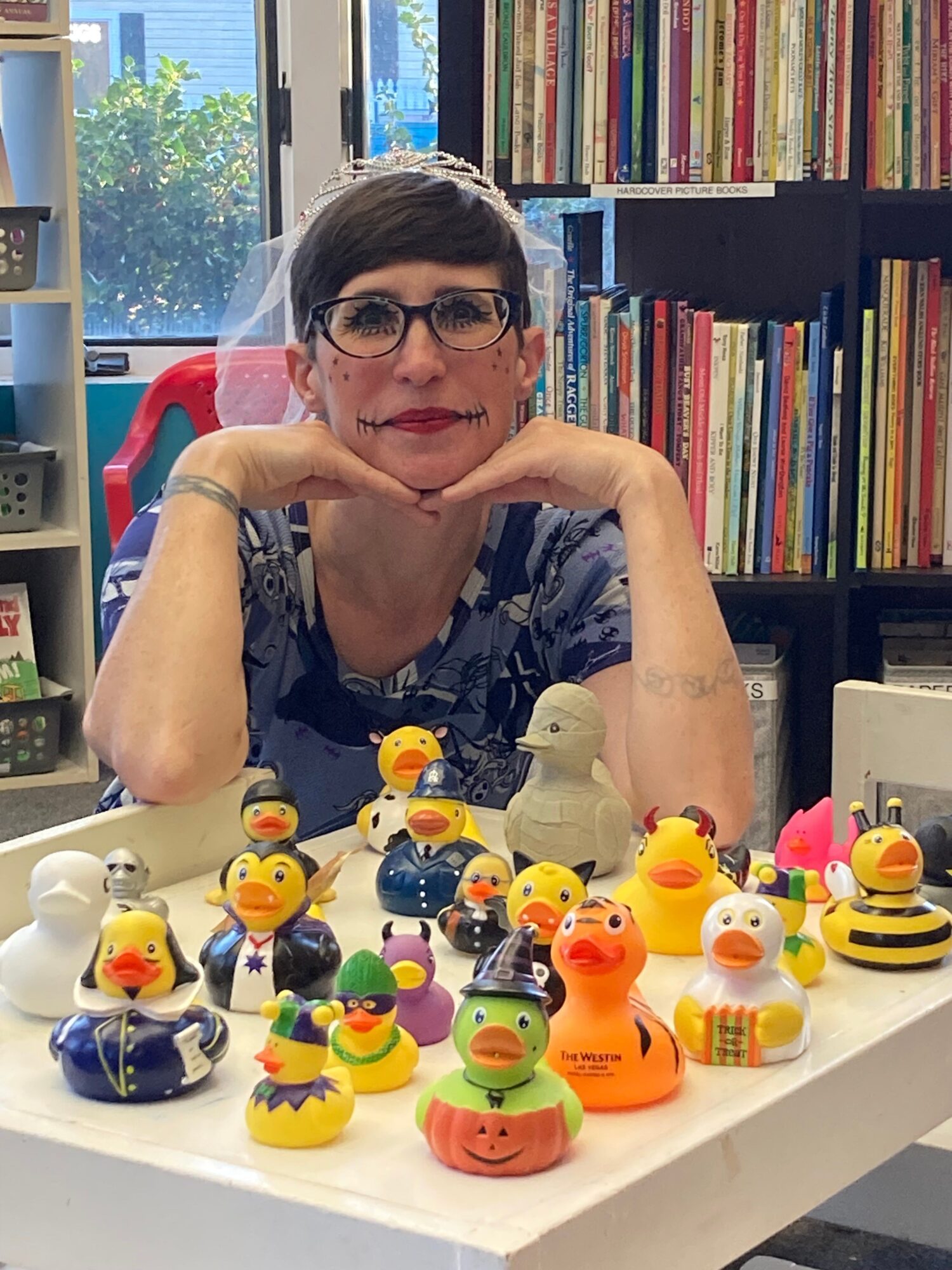 Woman with glasses and dark hair, smiling, surrounded by colorful rubber duck toys on a table, bookshelves in background.