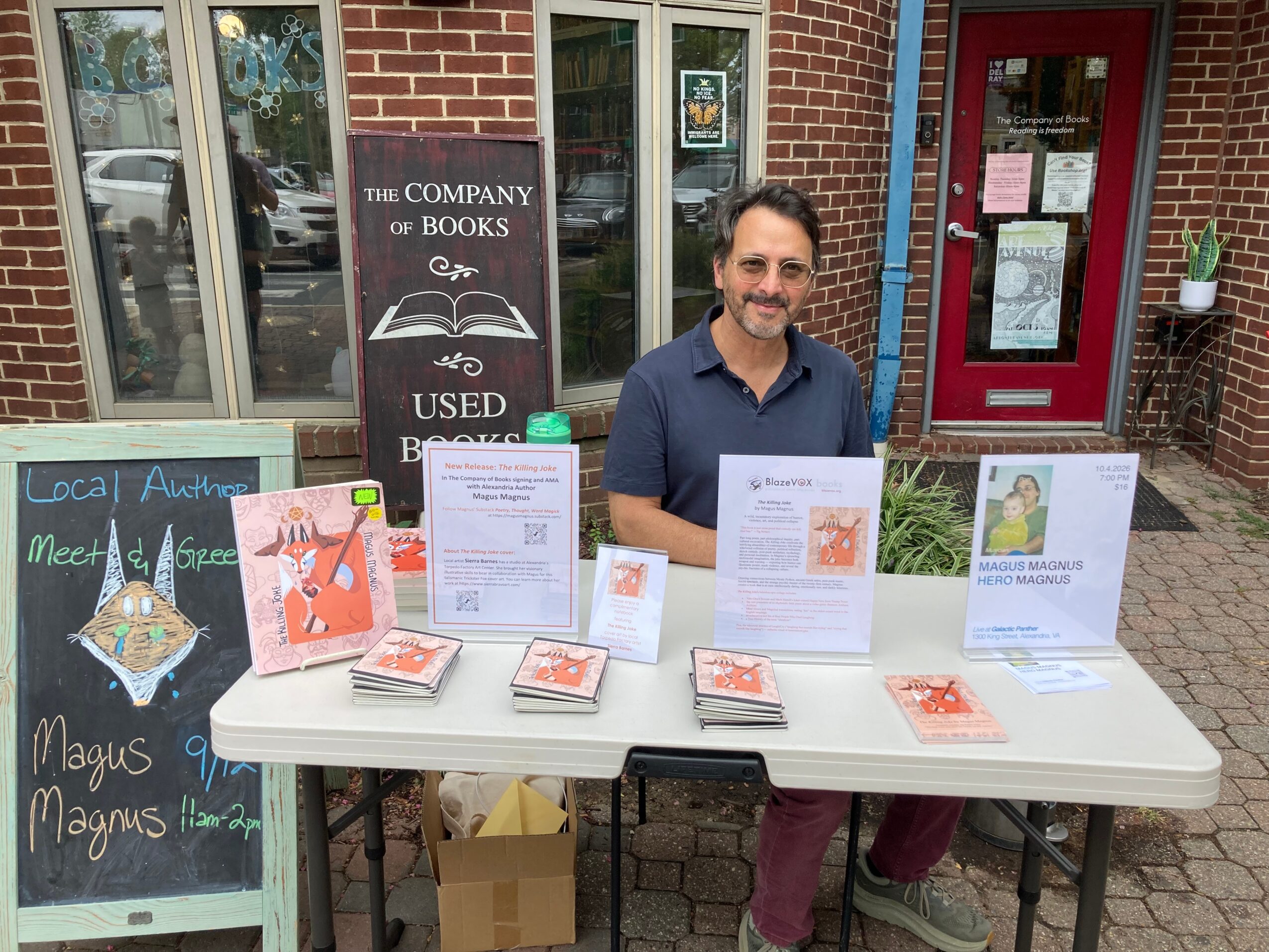Man standing behind a table with books and signs outside a brick building, with a window and red door in the background.