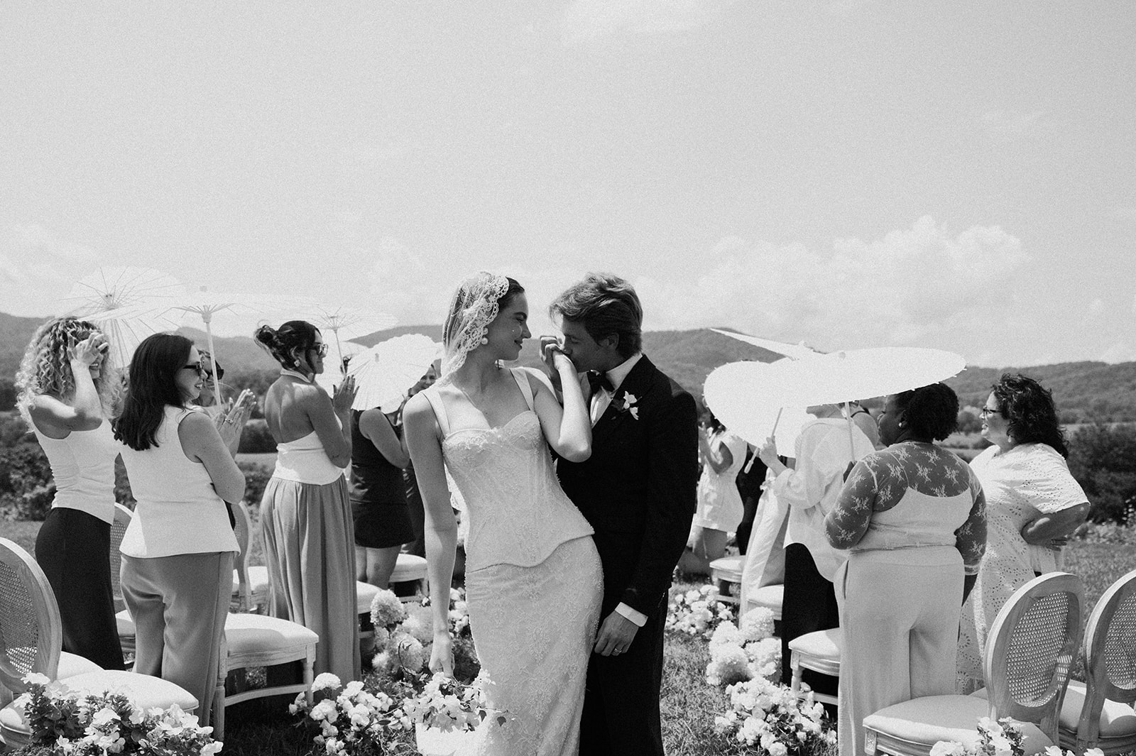 A wedding ceremony with a bride and groom standing close, surrounded by guests outdoors, with chairs and umbrellas, in black and white.