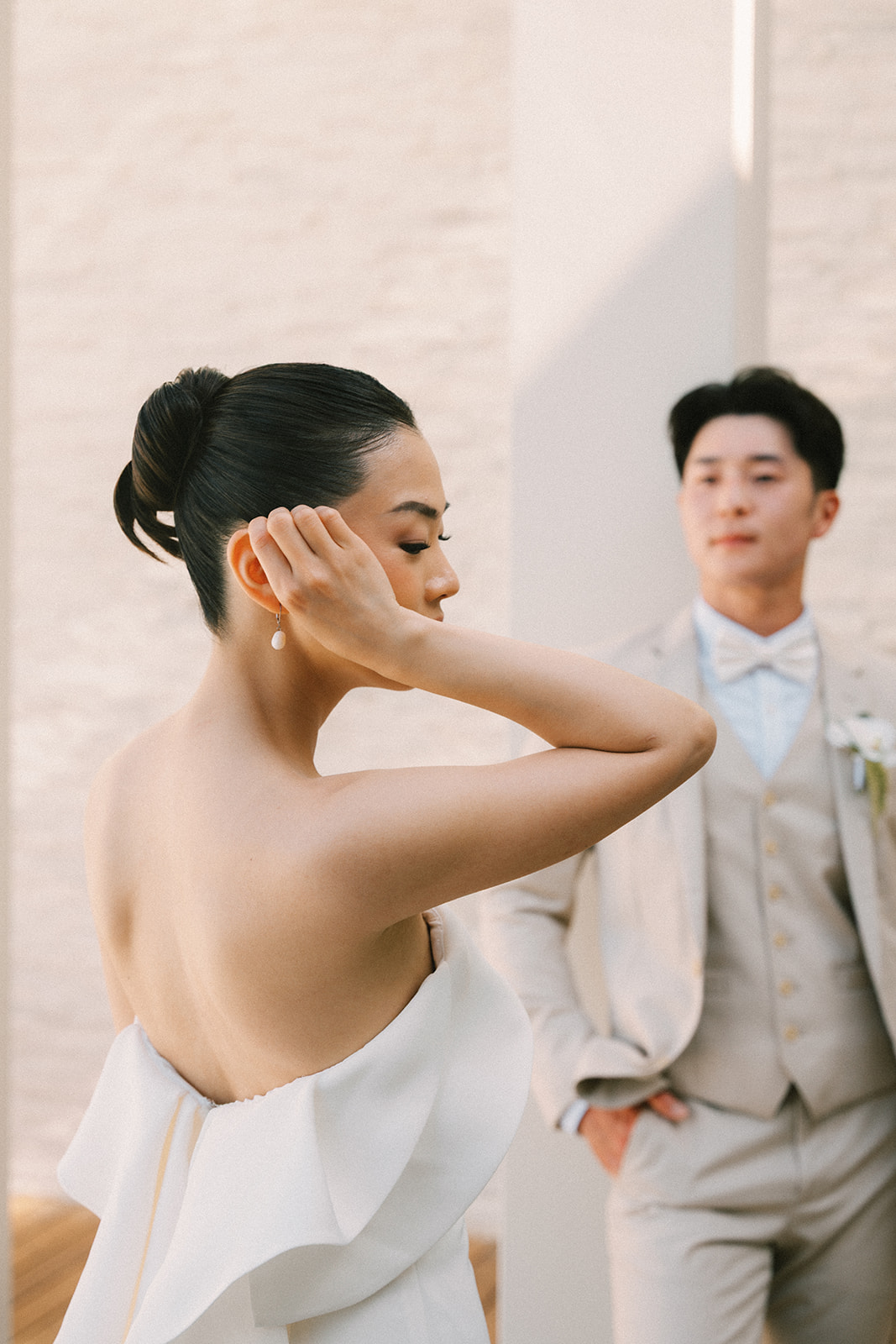 A woman with dark hair in a bun, wearing a strapless white dress, is adjusting her earring. A man in a light suit stands in the background.