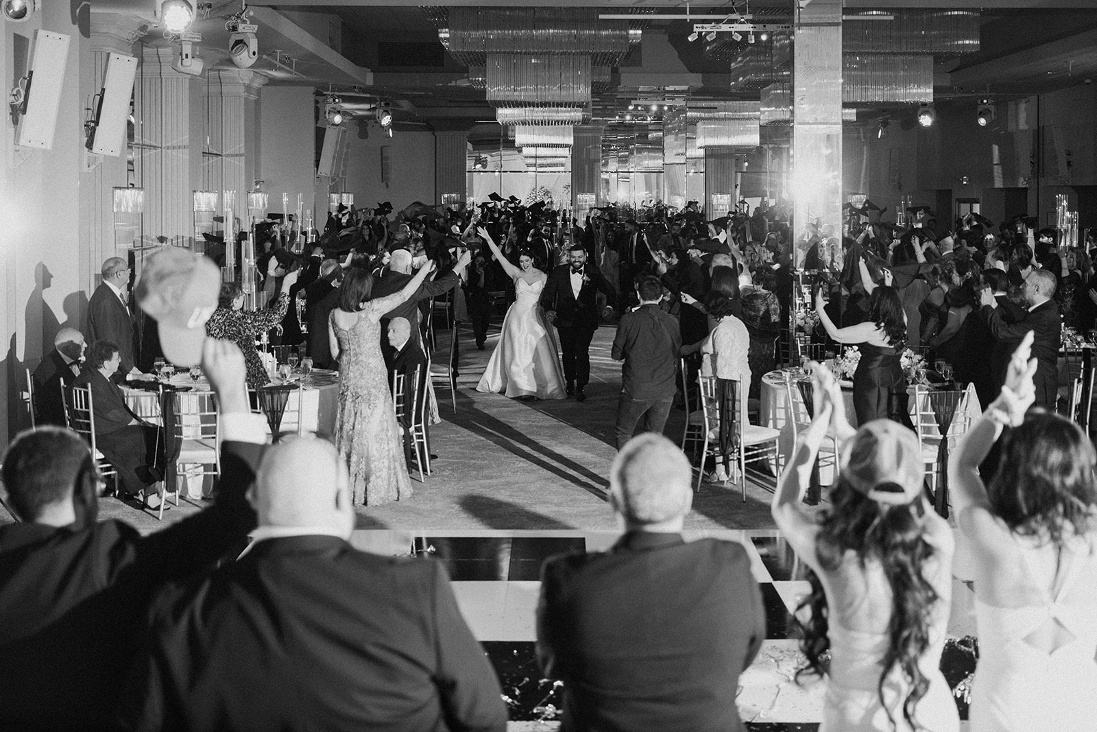 Crowd of people dancing and socializing in a large banquet hall with tables and chandeliers.