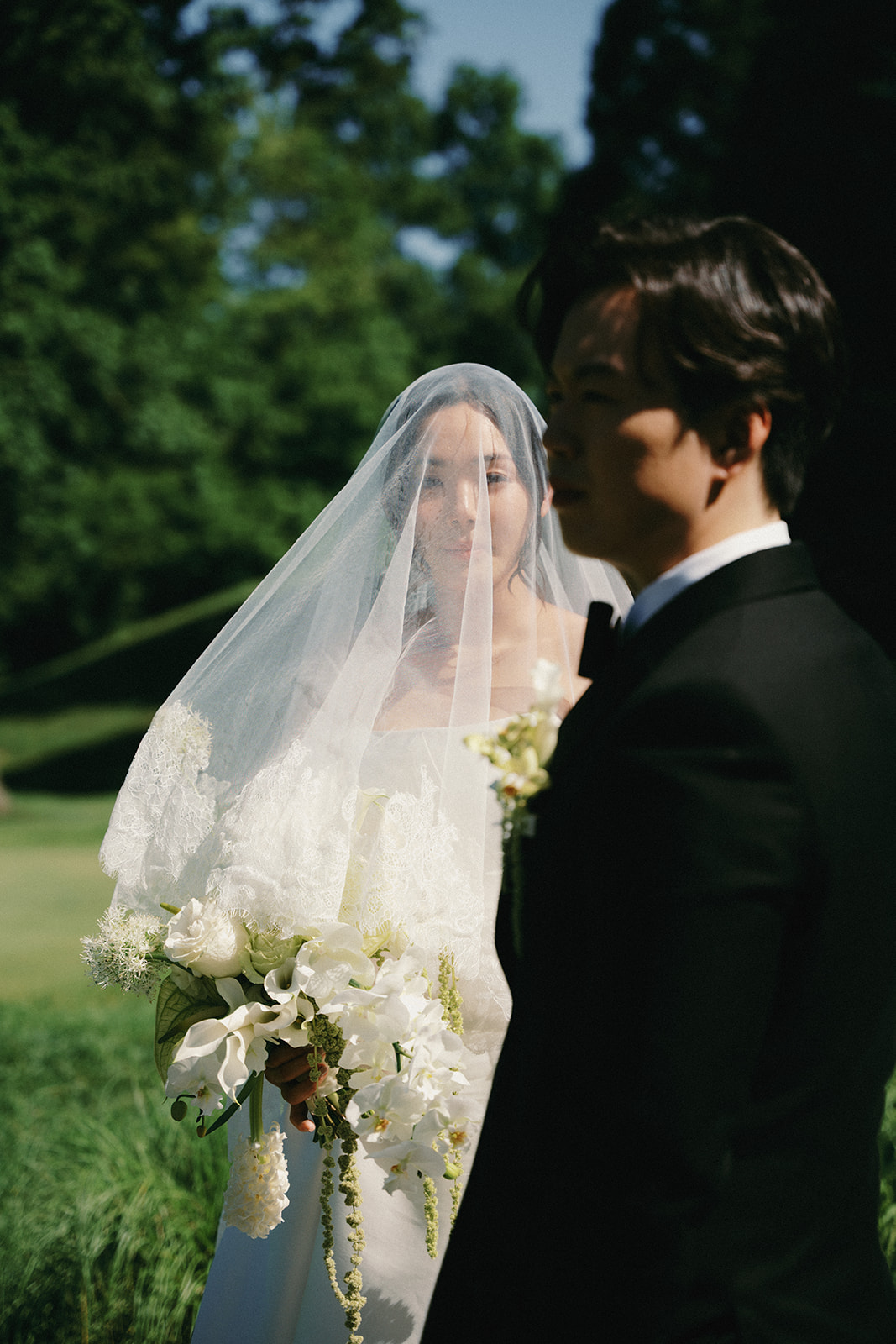 Bride in wedding dress with veil holding bouquet, groom in tuxedo outdoors with trees in background.