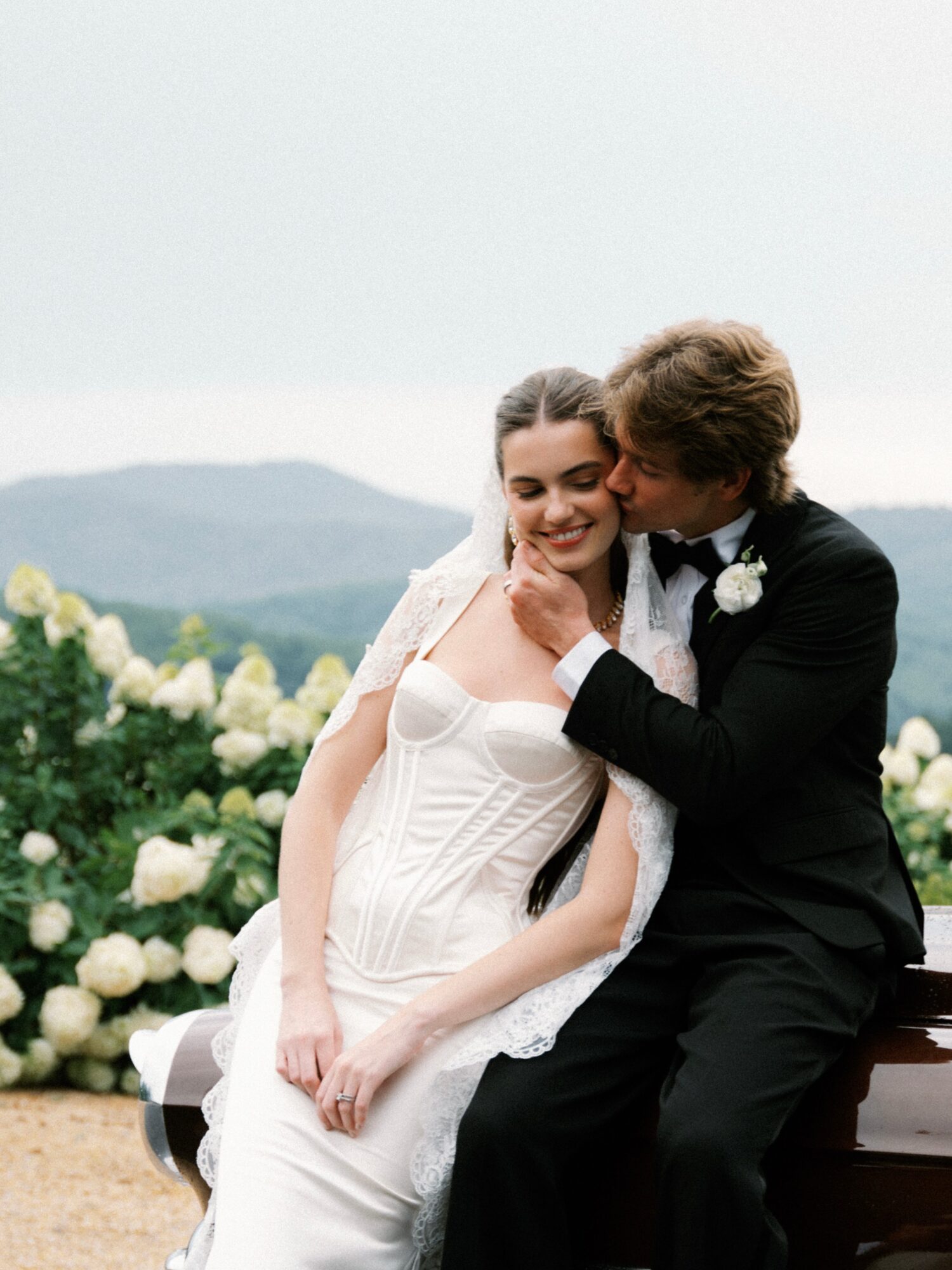 A couple sits on a bench outdoors, with mountains and white flowers in the background, sharing a kiss and smiling.