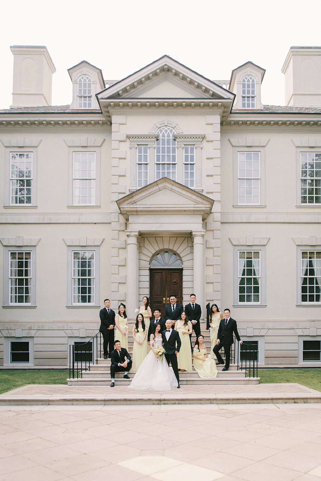 Group of people in formal attire standing on steps in front of a large, elegant mansion with multiple windows and a central entrance.
