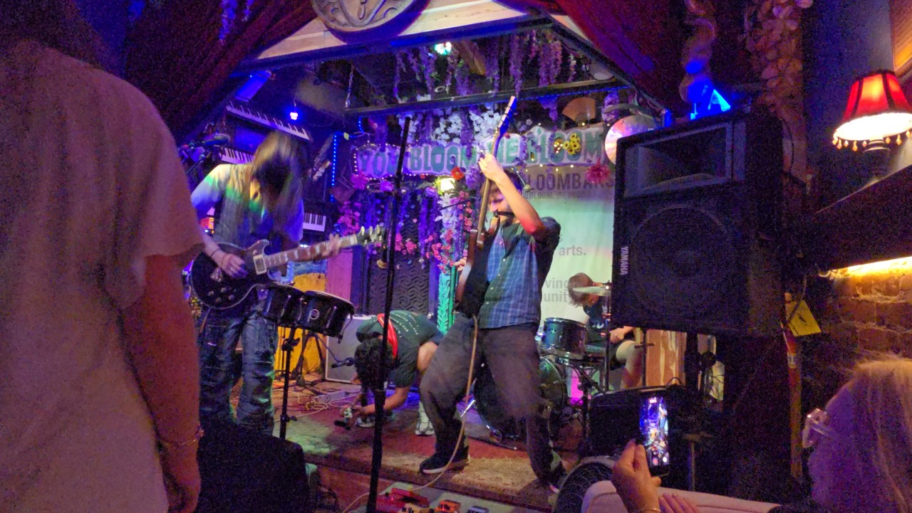 People performing on a small stage in a dimly lit venue with colorful lights and musical instruments.