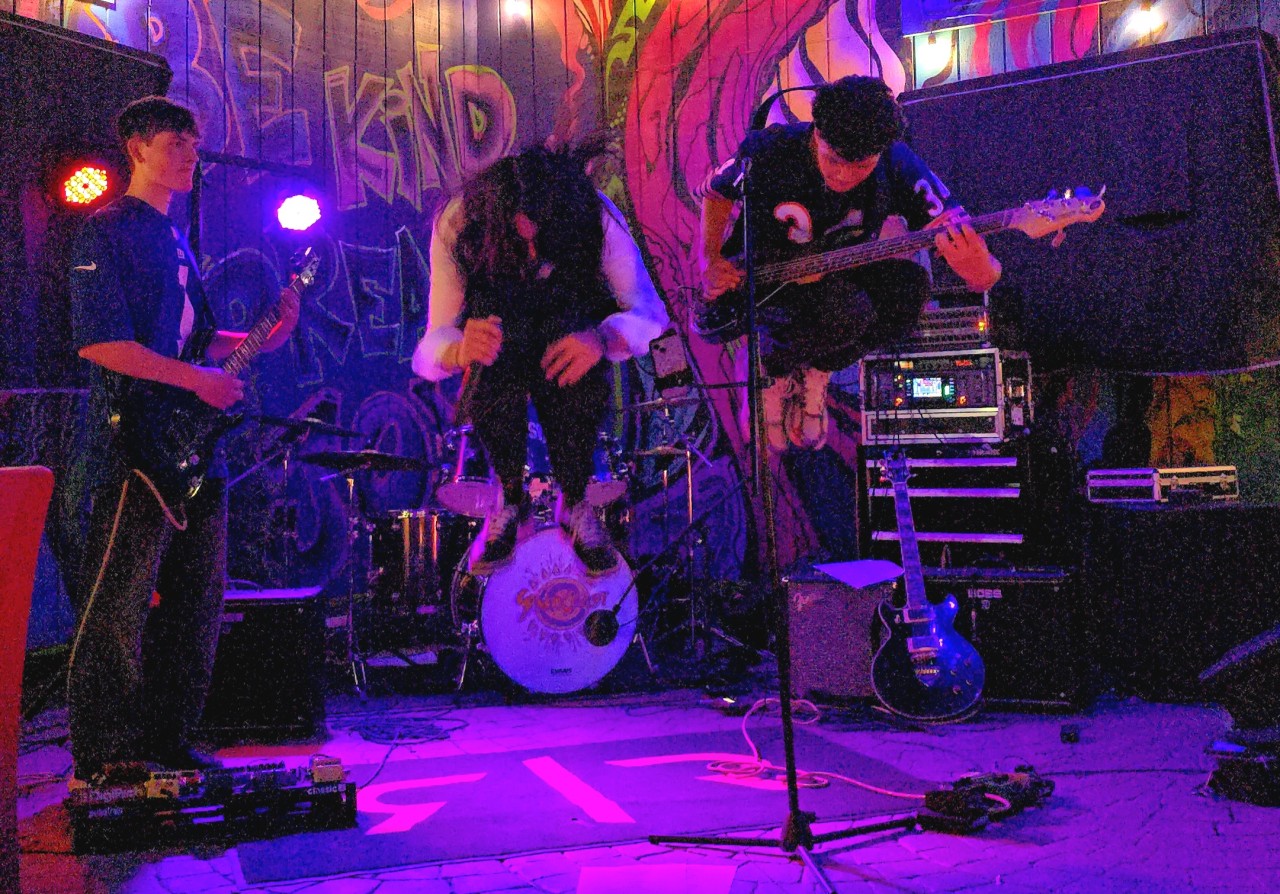 Three musicians perform on stage with colorful graffiti background, playing guitars and singing, with musical equipment around them.