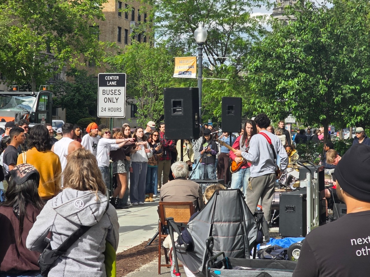 Crowd gathered around a speaker system in a park with trees and buildings in the background.