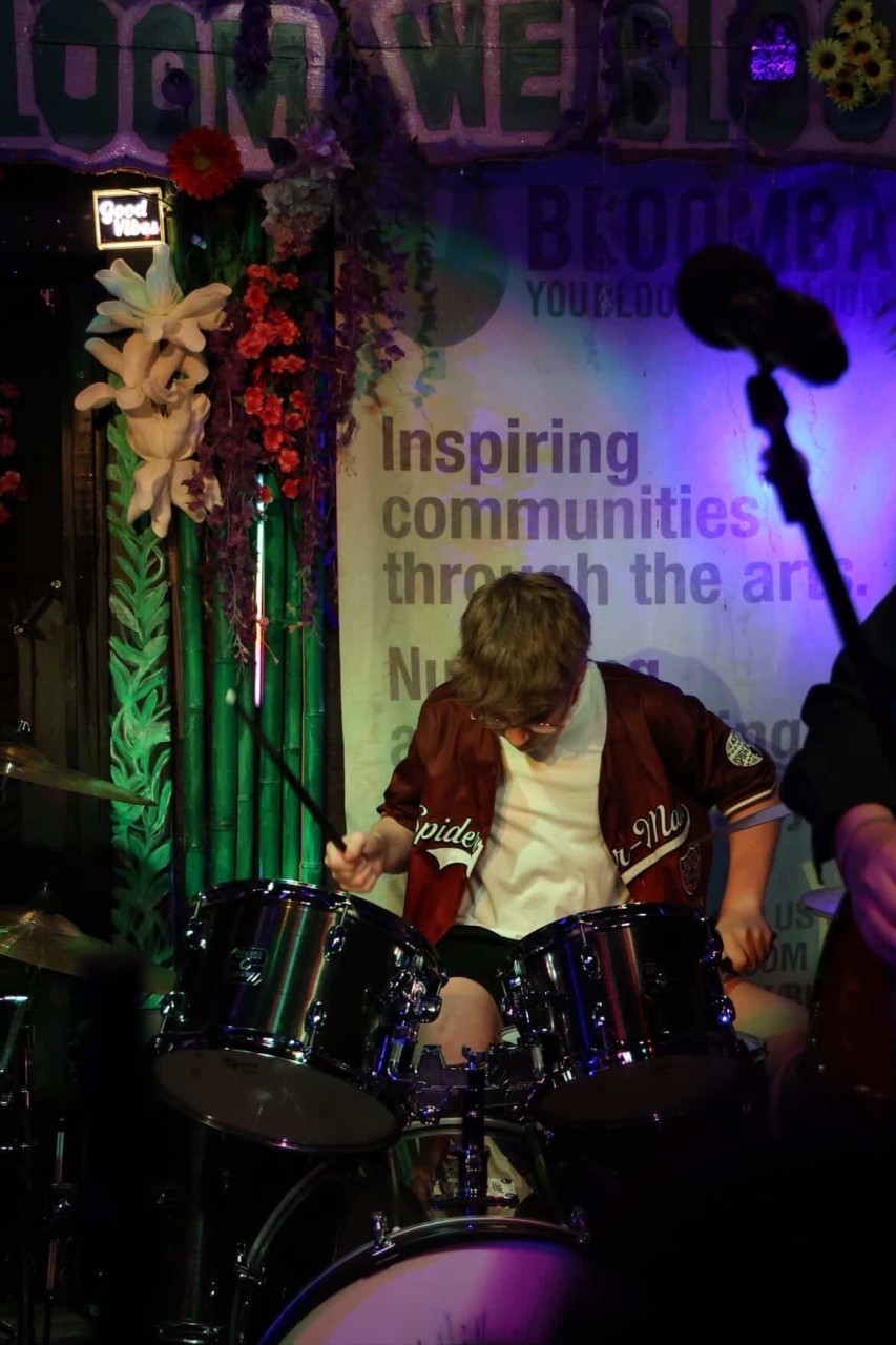 Child playing drums on stage with colorful decorations and a microphone in the background.