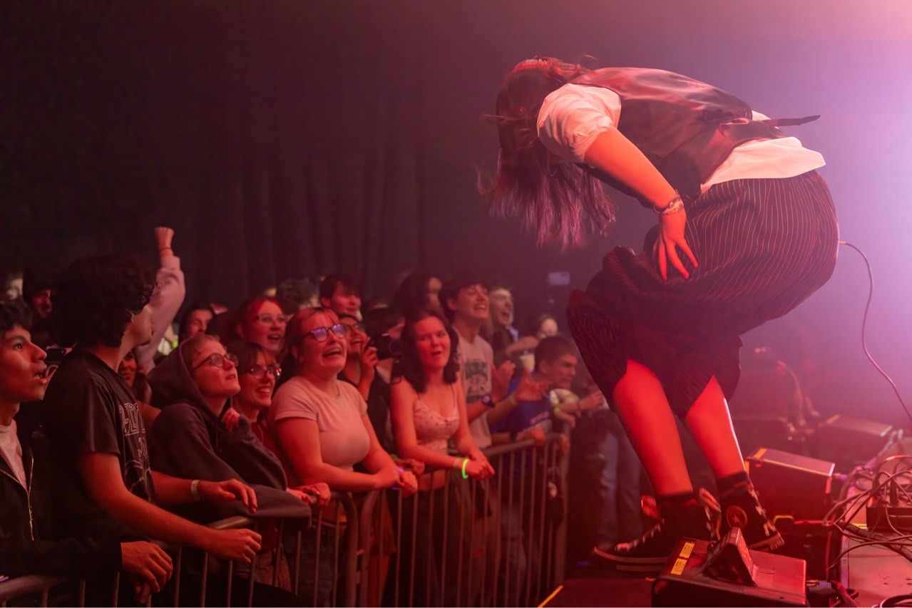 Performer on stage bending forward with hair covering face, audience watching attentively, stage lights in background.