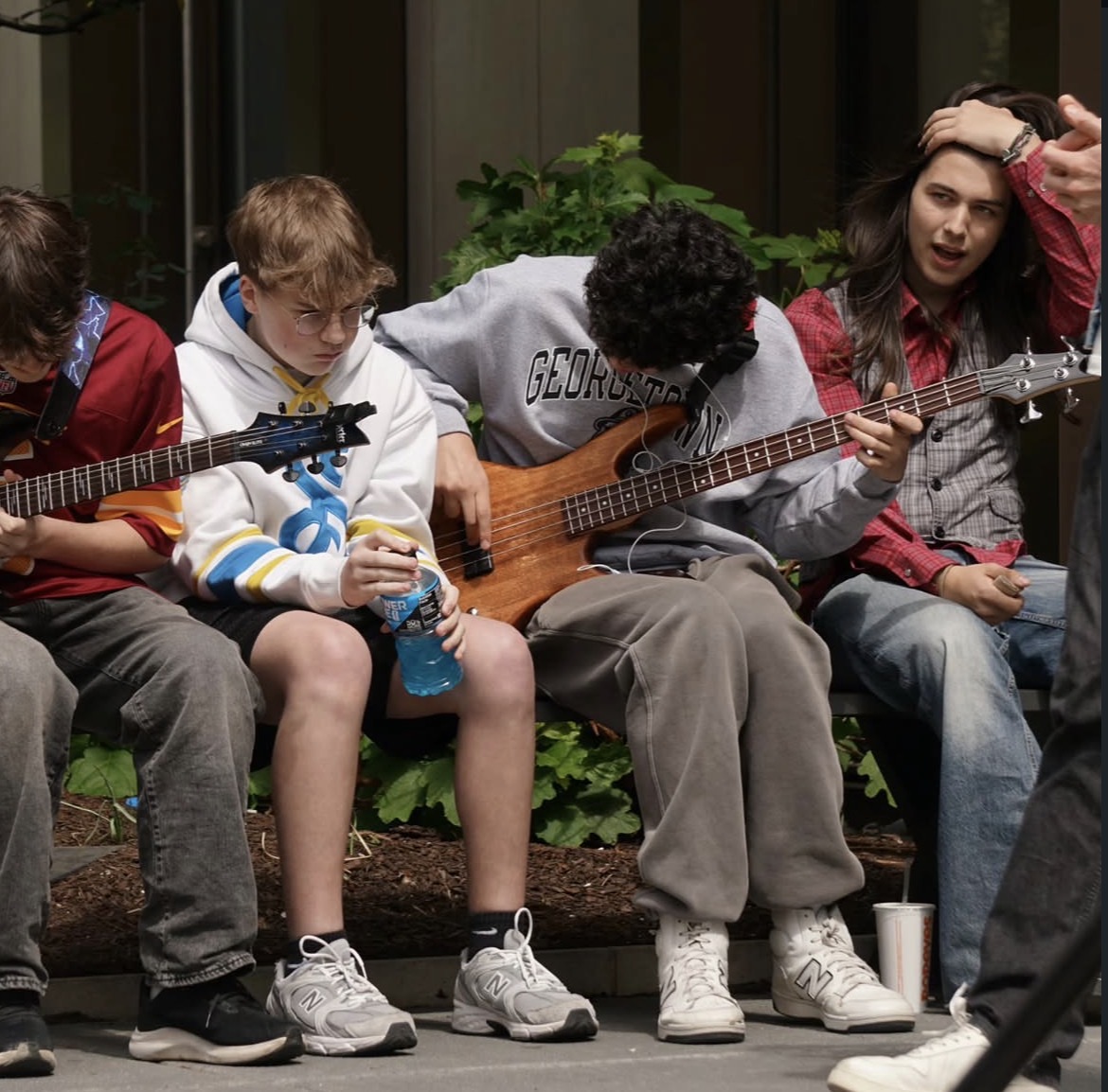 Group of young people sitting outdoors, some holding guitars, one person drinking from a bottle, surrounded by greenery.