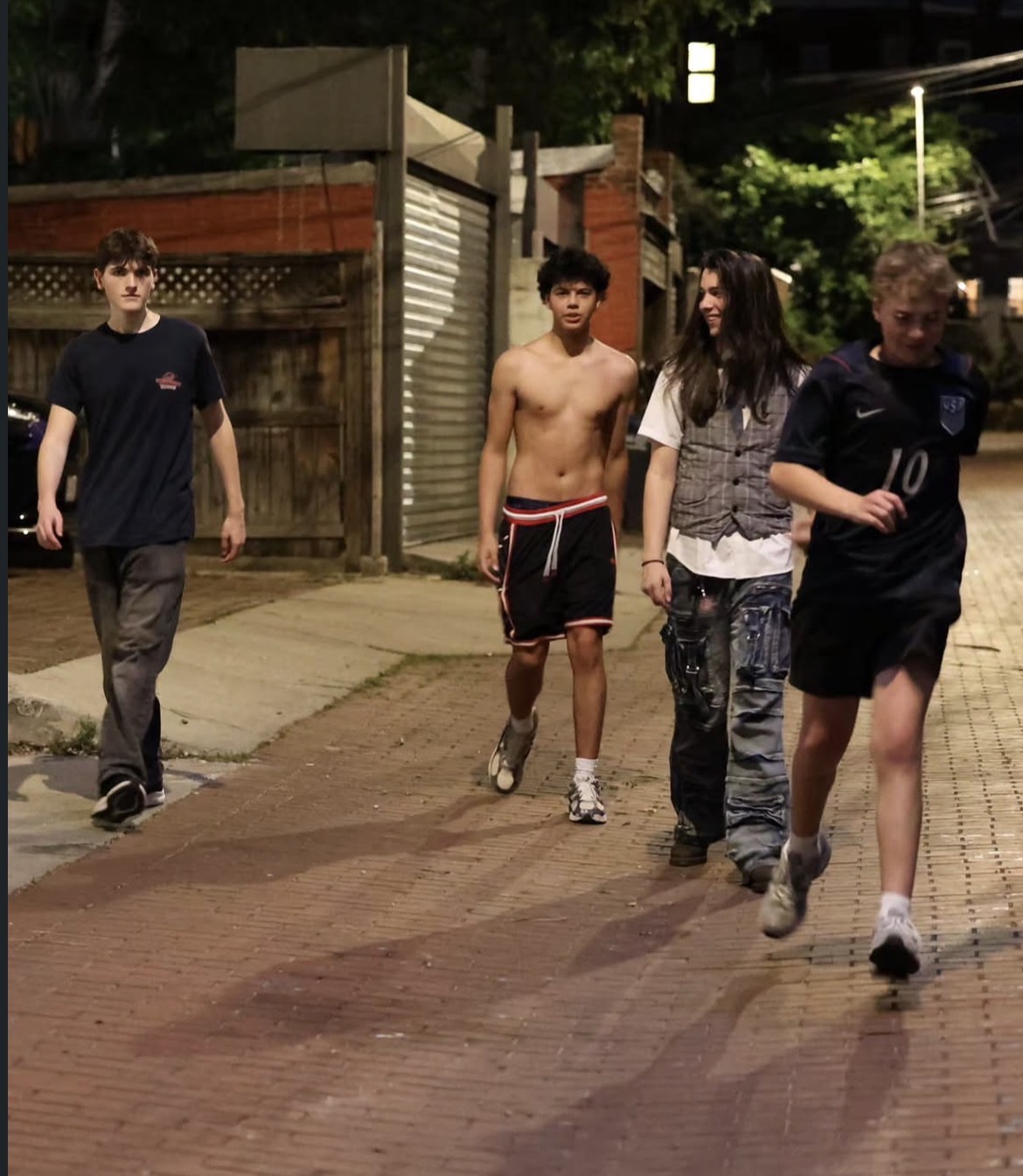 Four young people walking on a sidewalk at night, some wearing casual clothes, one shirtless, with buildings and trees in the background.