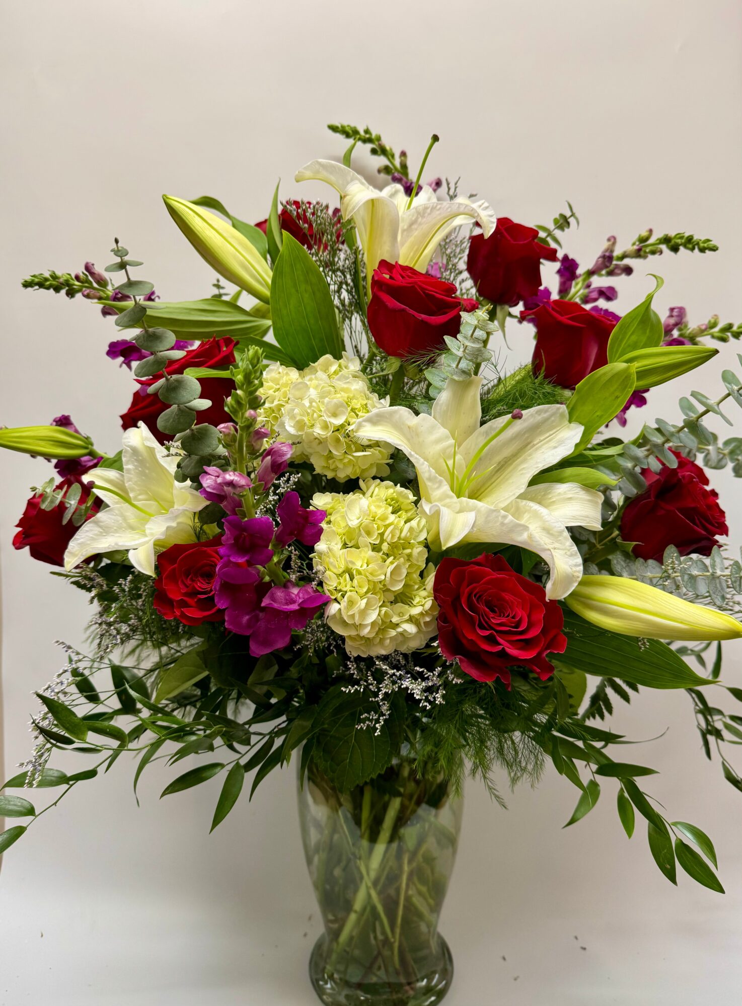 A bouquet of white lilies, red roses, purple flowers, and green foliage in a glass vase.