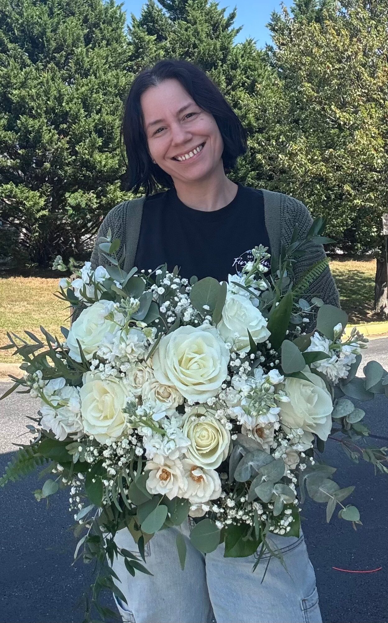 Person with short dark hair smiling outdoors holding a large bouquet of white flowers, trees in background.