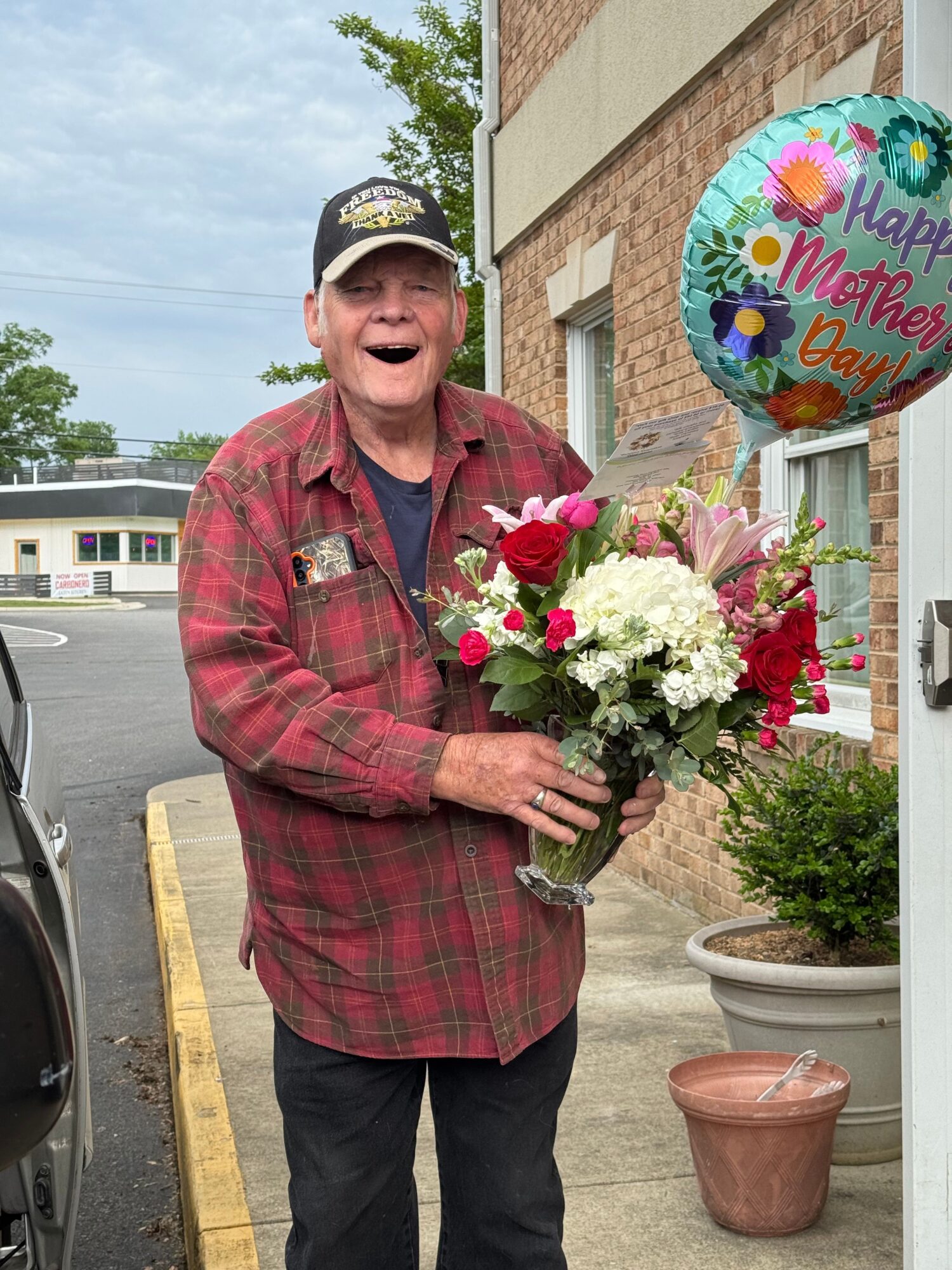 Person holding a large bouquet of flowers, wearing a plaid jacket and cap, standing outdoors near a building.