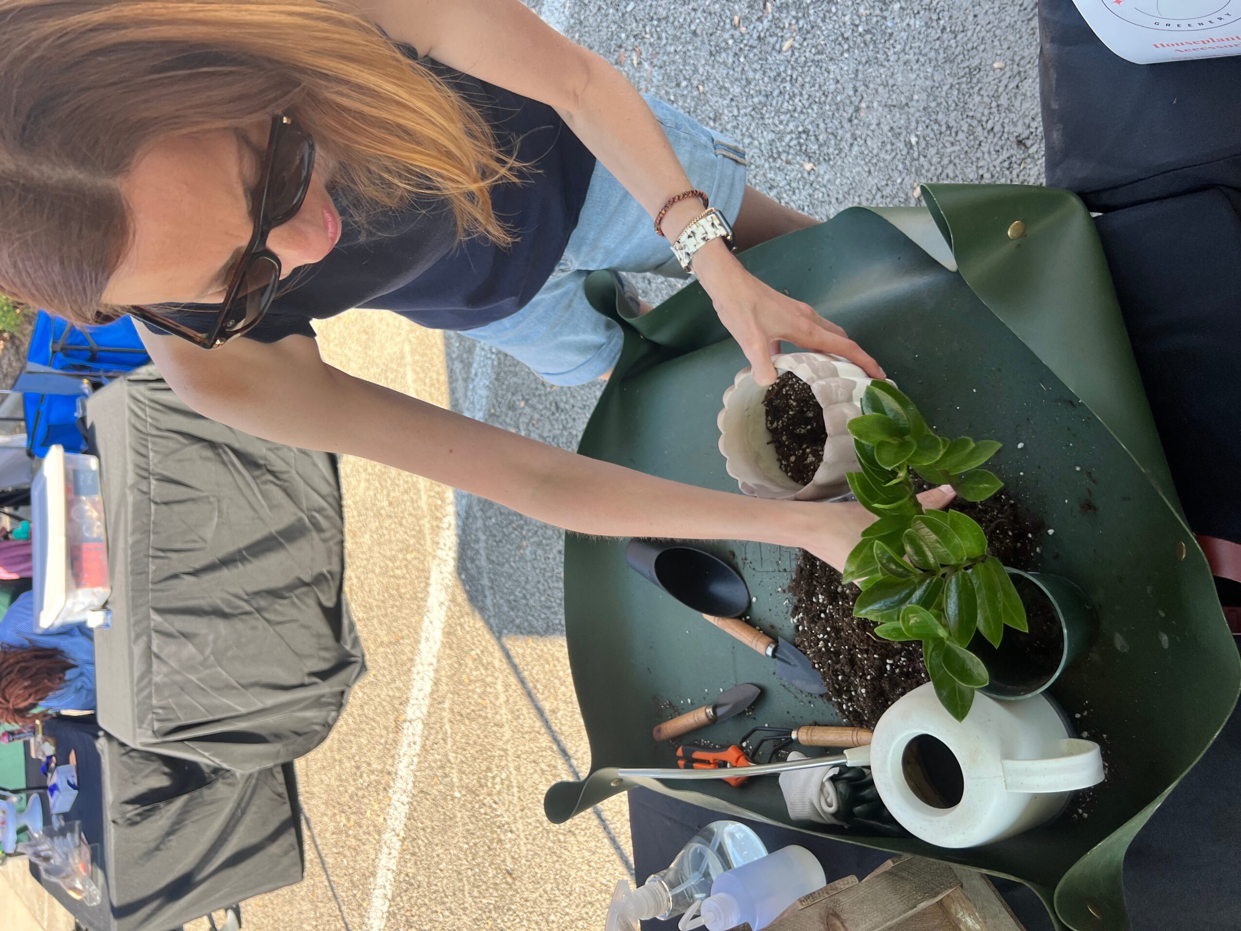 Person planting a small green plant in soil on a green surface outdoors, with gardening tools nearby.