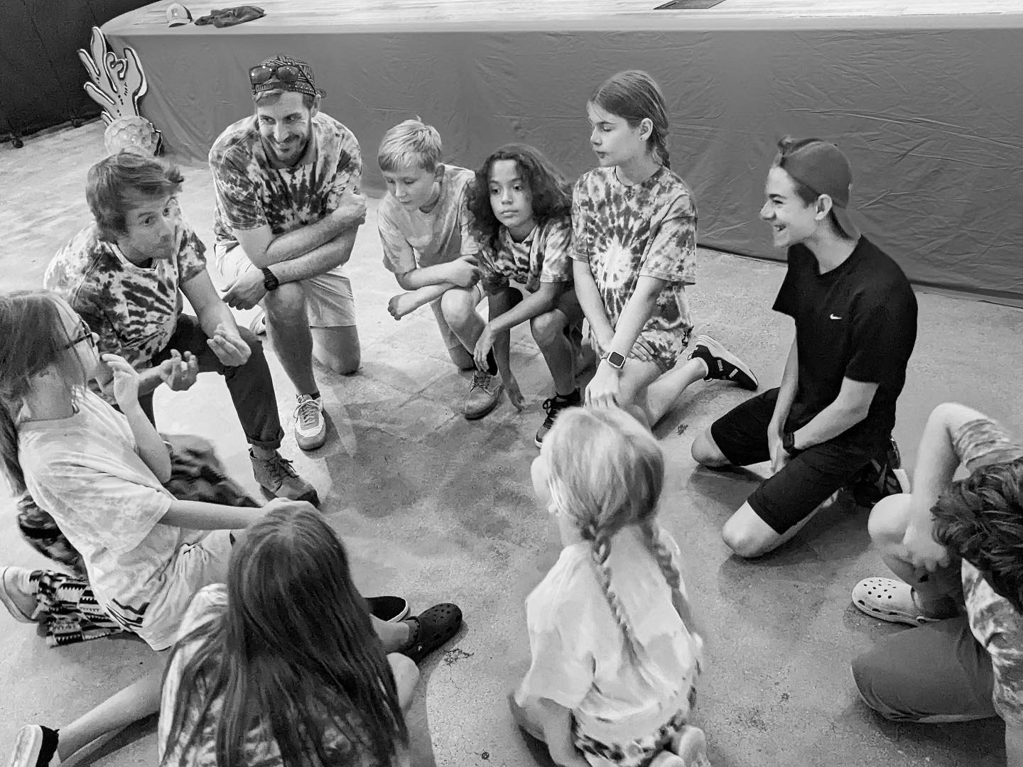Children and an adult sit in a circle on the floor, engaging in a group activity indoors.