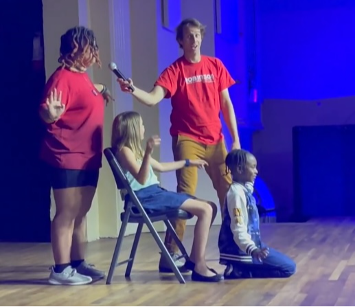 Two women and two children on stage, one woman holding a microphone, children sitting and kneeling, blue background, wooden floor.