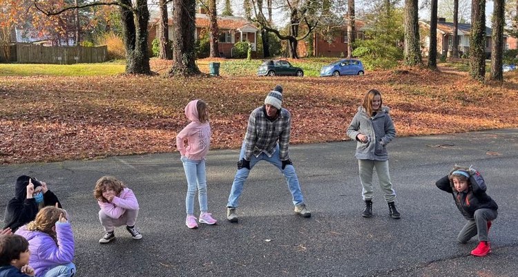 Four children standing on a paved surface outdoors, with trees and parked cars in the background.