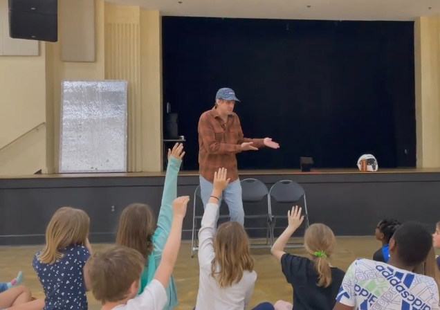 Person on stage with children raising hands in audience, stage with black backdrop, children seated and engaging.