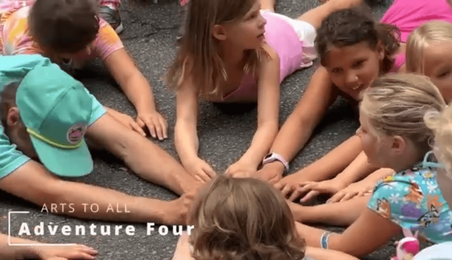 Group of children lying on the ground with hands stacked together, smiling and engaging in an outdoor activity.