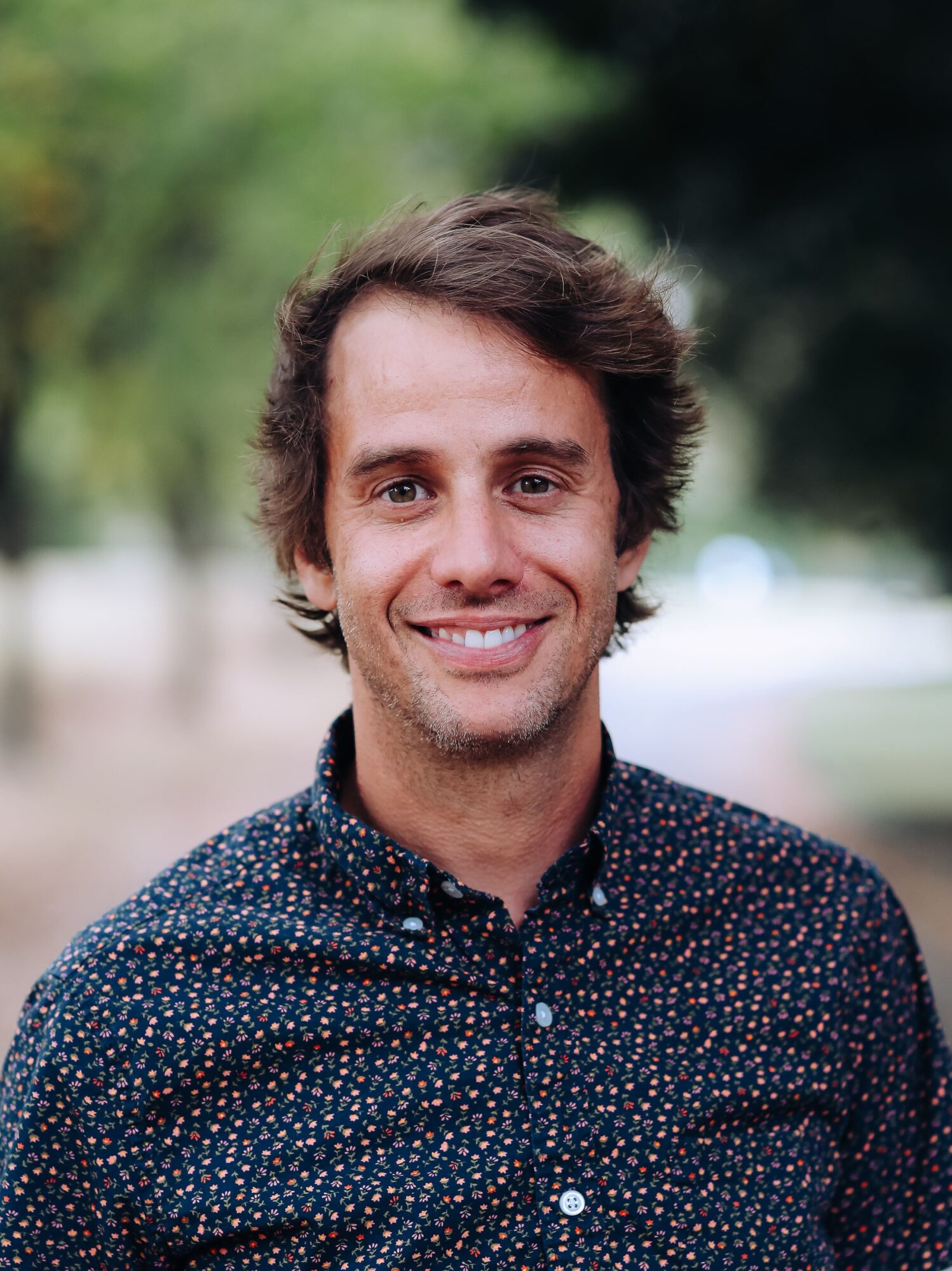 Young man with curly brown hair smiling outdoors, wearing a dark patterned shirt, blurred green background.