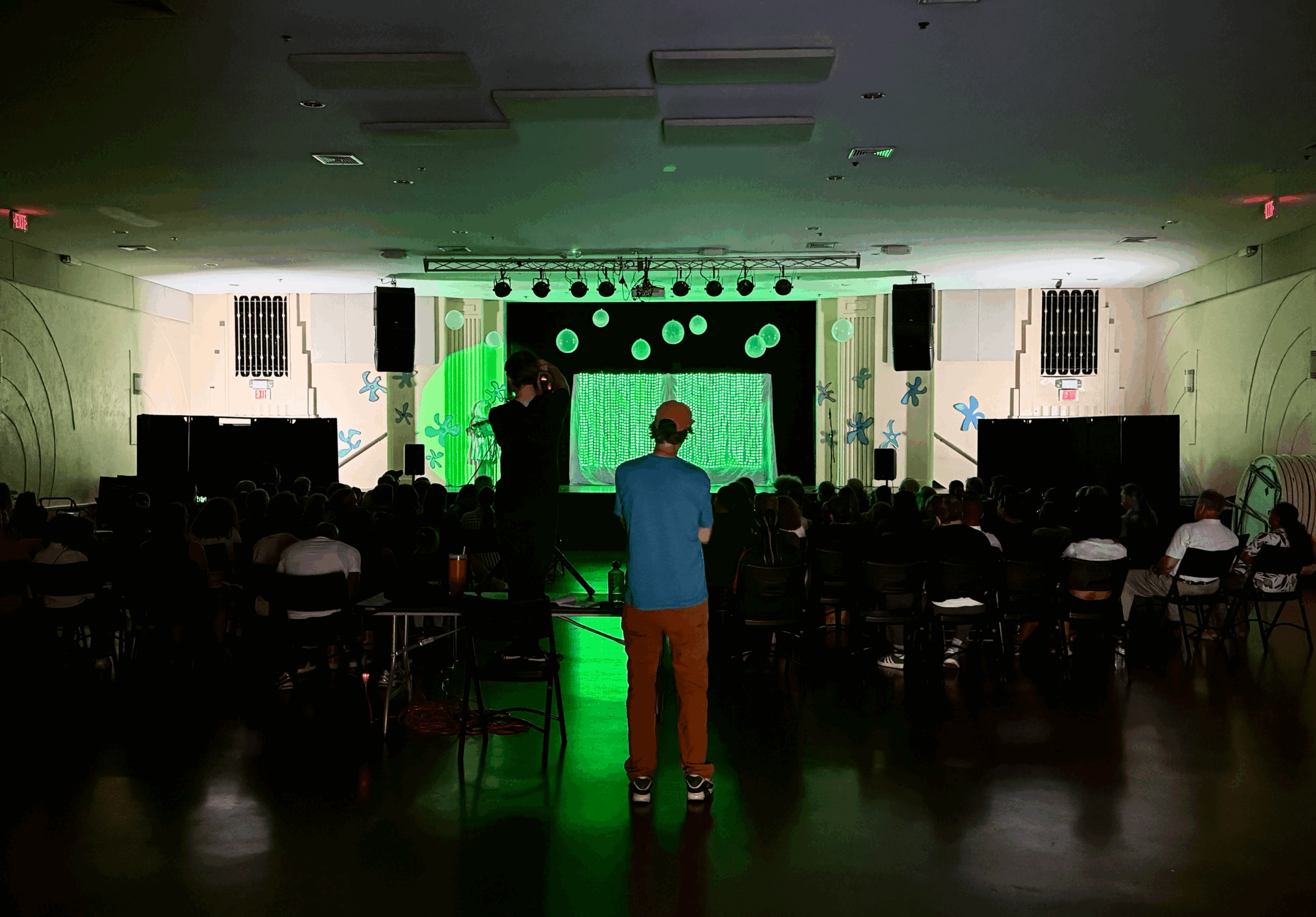 Person standing in front of a stage with a large screen, audience seated in a dark room, stage lit with green and white lights.