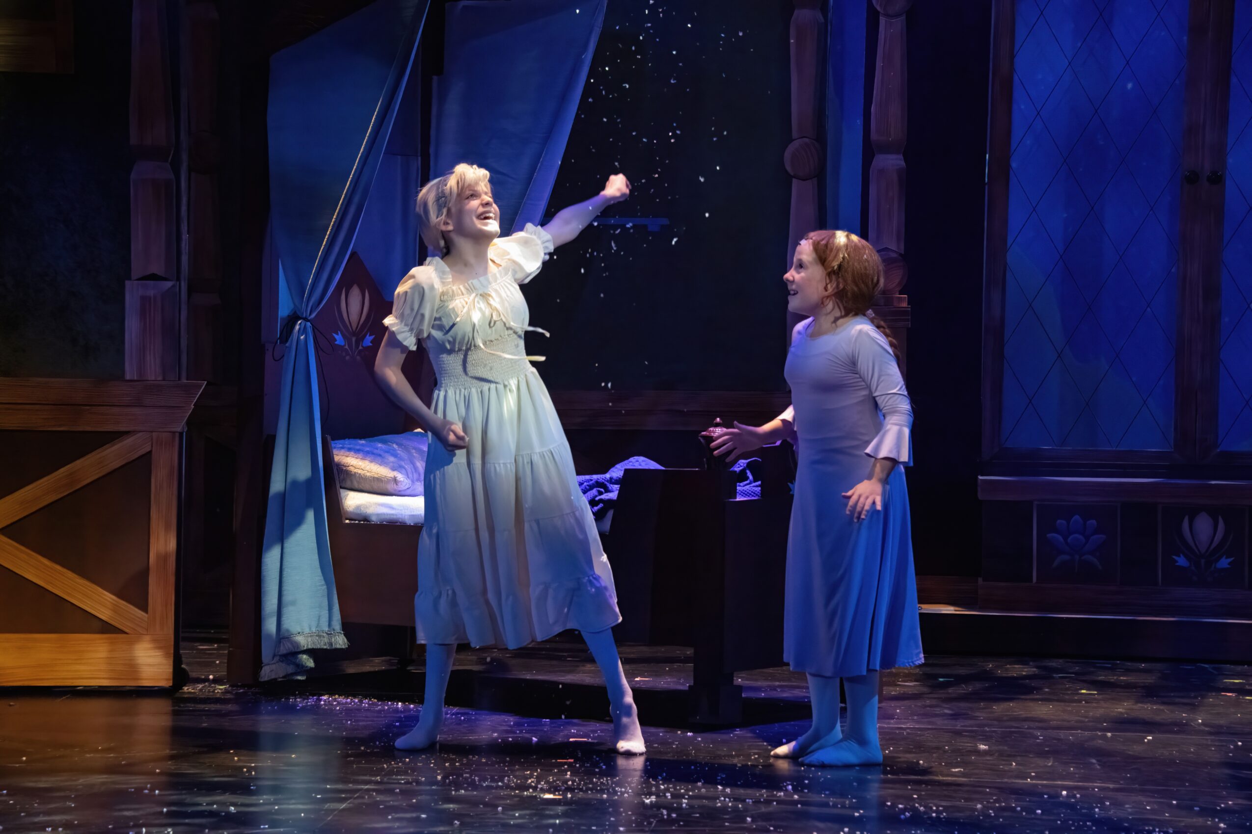 Two women on stage, one reaching out, with a curtain and stage set in the background.