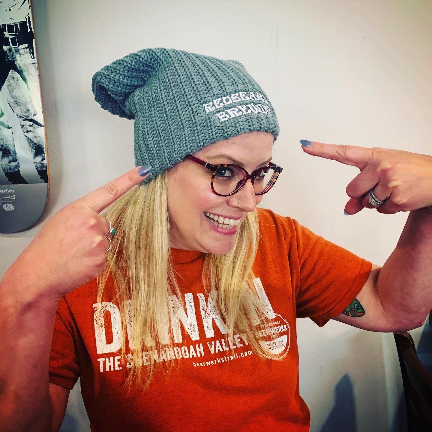 Woman wearing glasses, a gray beanie, and an orange shirt, smiling and pointing at her head.